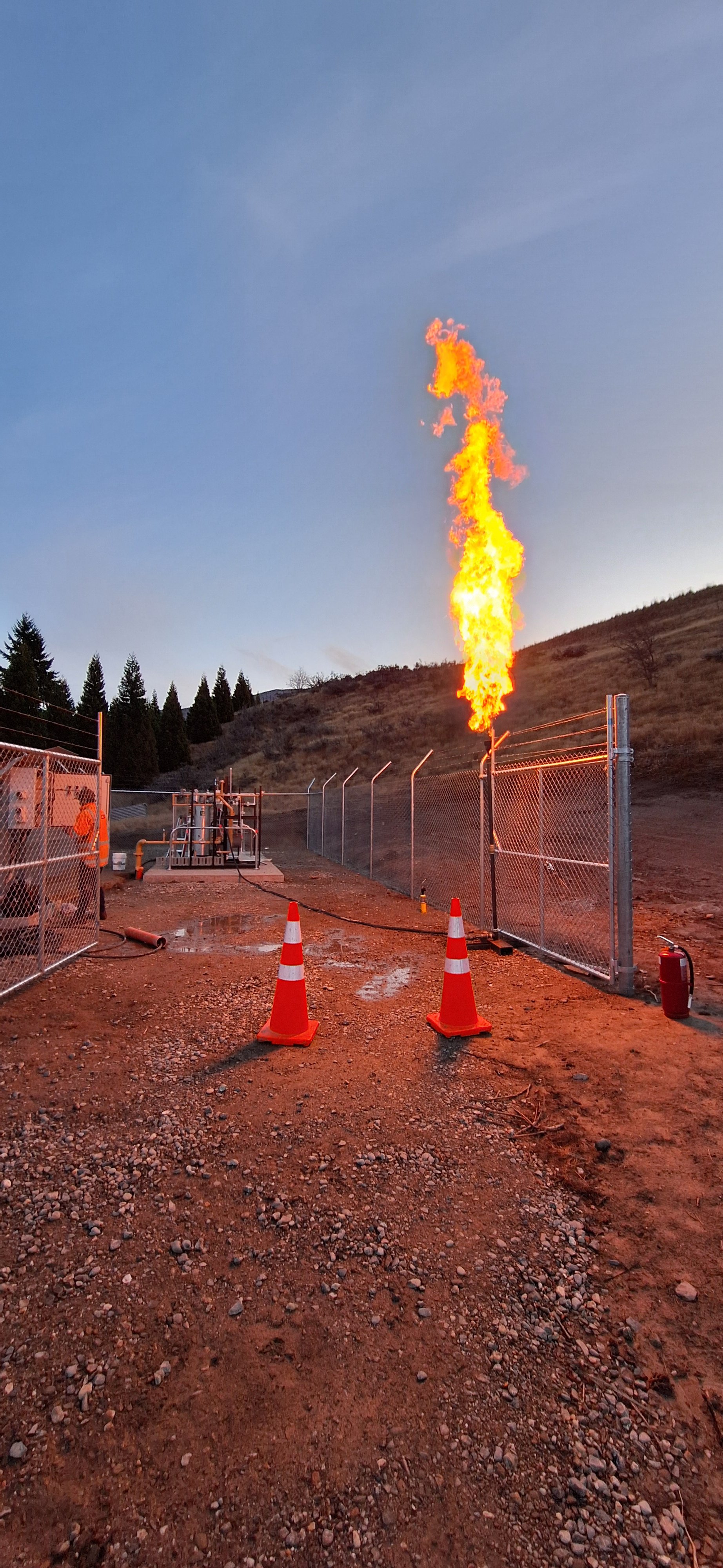 A gas flare on a pipeline site with a flame shooting upward, enclosed by chain-link fencing, in a rural area with hills and trees in the background.