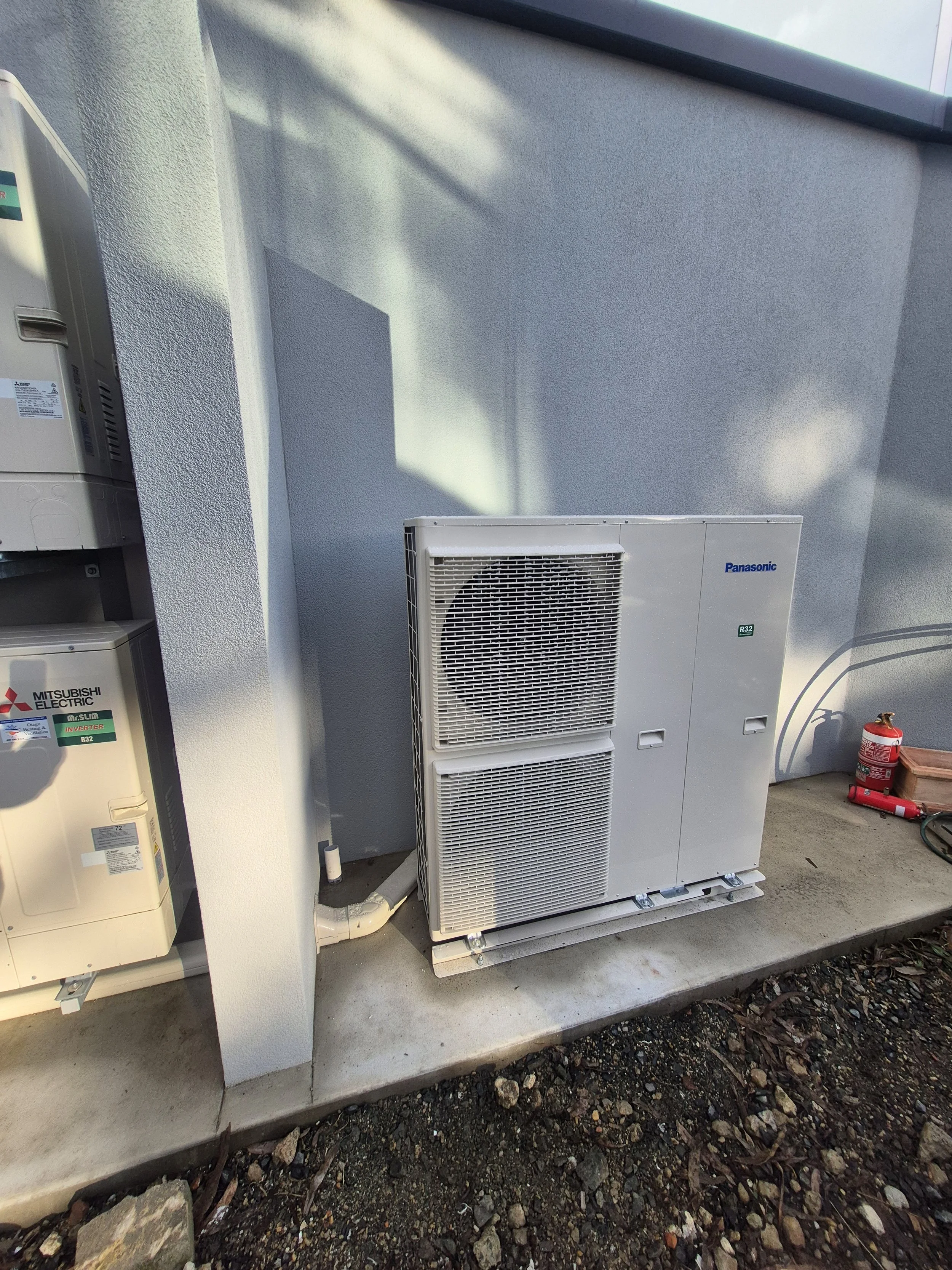 Outdoor view of HVAC equipment including an air conditioning unit and electrical boxes mounted on a concrete pad against the exterior wall of a building