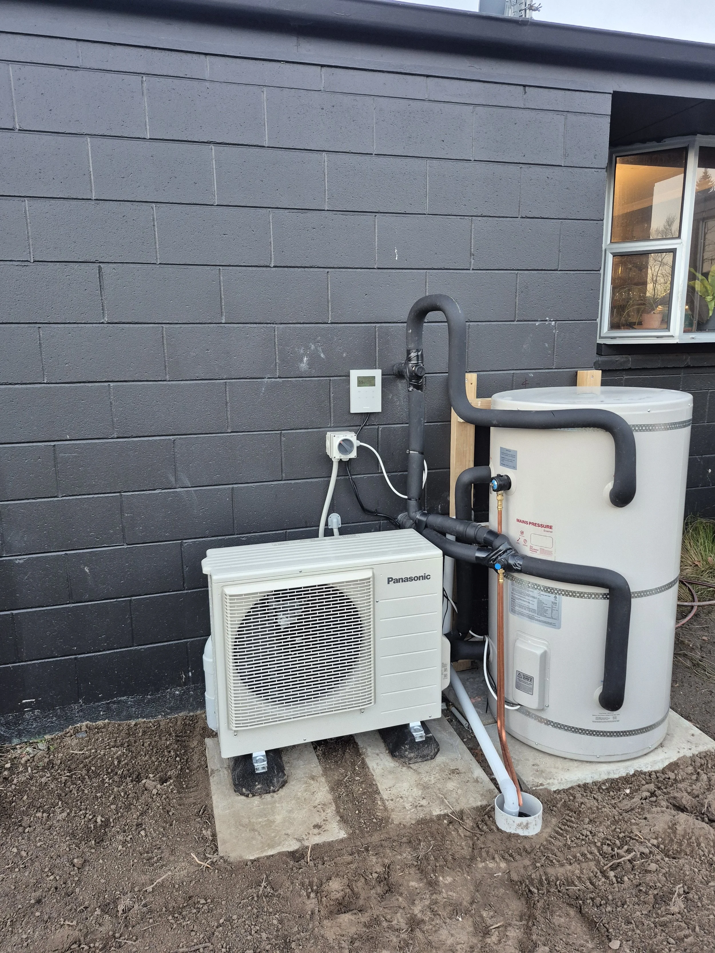 Outdoor HVAC system setup against a gray brick wall, including an air conditioning unit and a water heater connected by pipes, with a window showing plants inside.