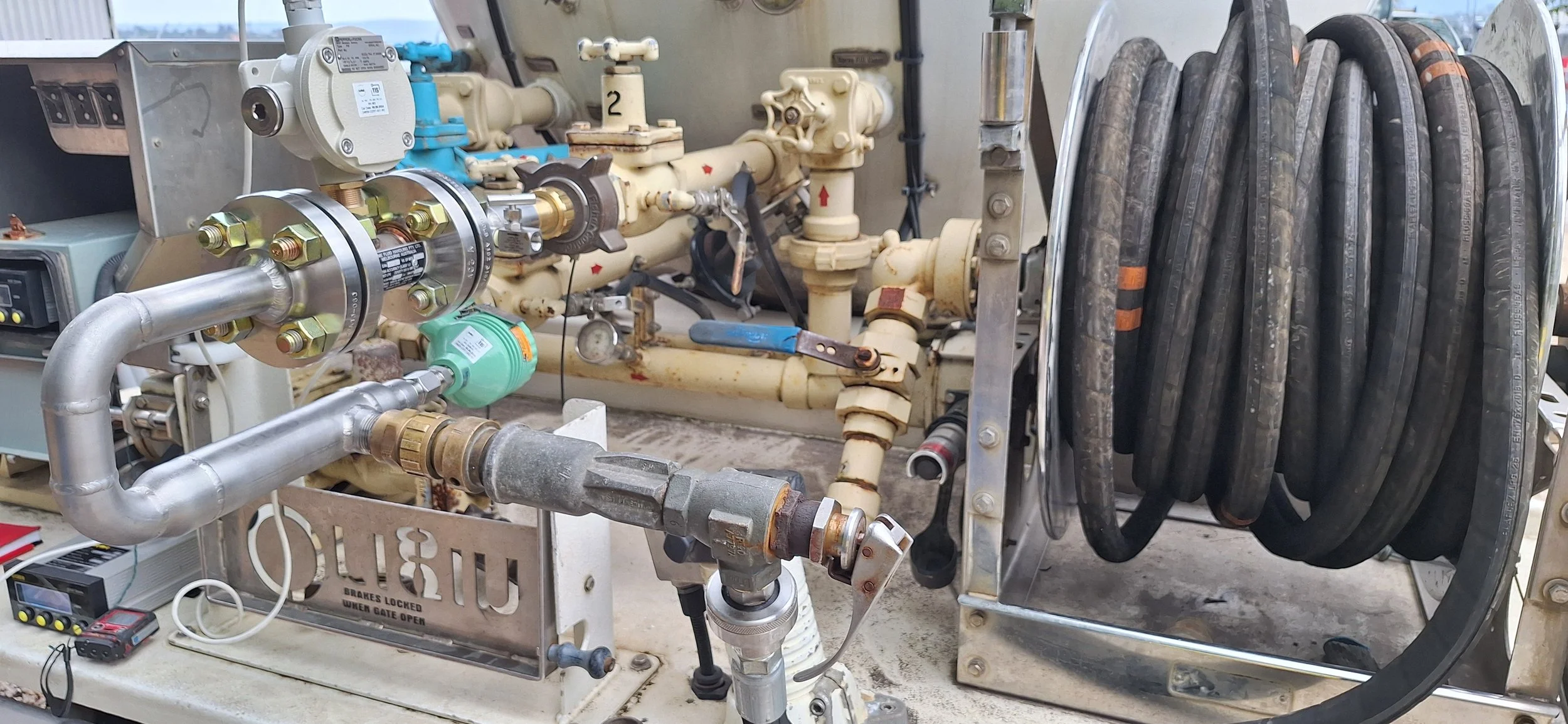 A collection of industrial pipes, valves, and hoses, including a large coiled hose on a metal reel, with additional equipment and tools in the background.