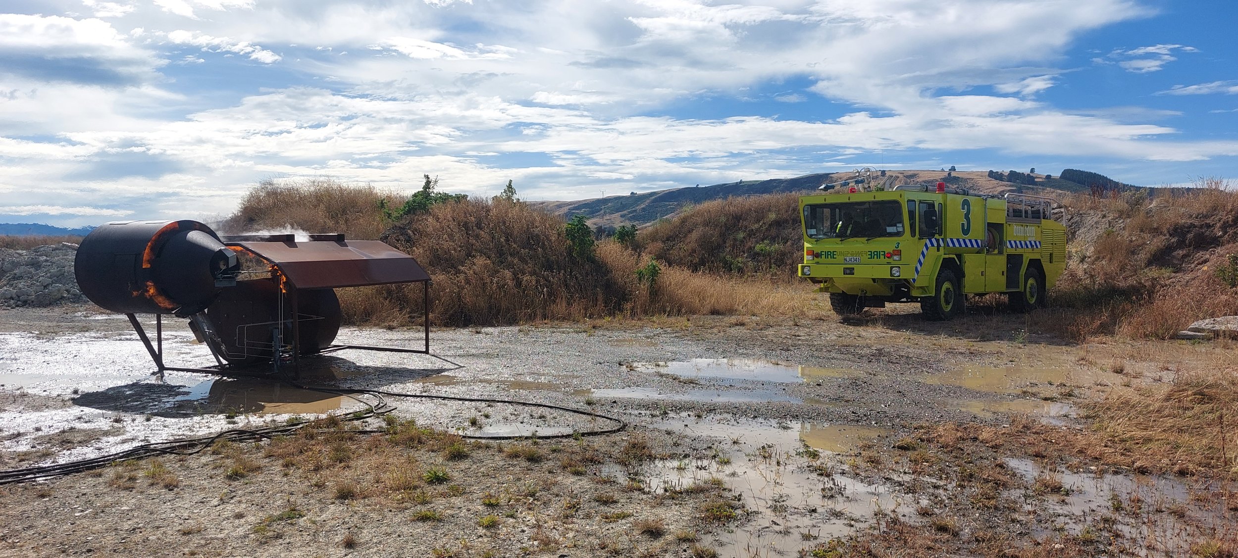 Firefighting scene with a flipped over barrel containing flammable liquid, surrounded by muddy water, with a yellow fire truck nearby on a dirt field under a partly cloudy sky.