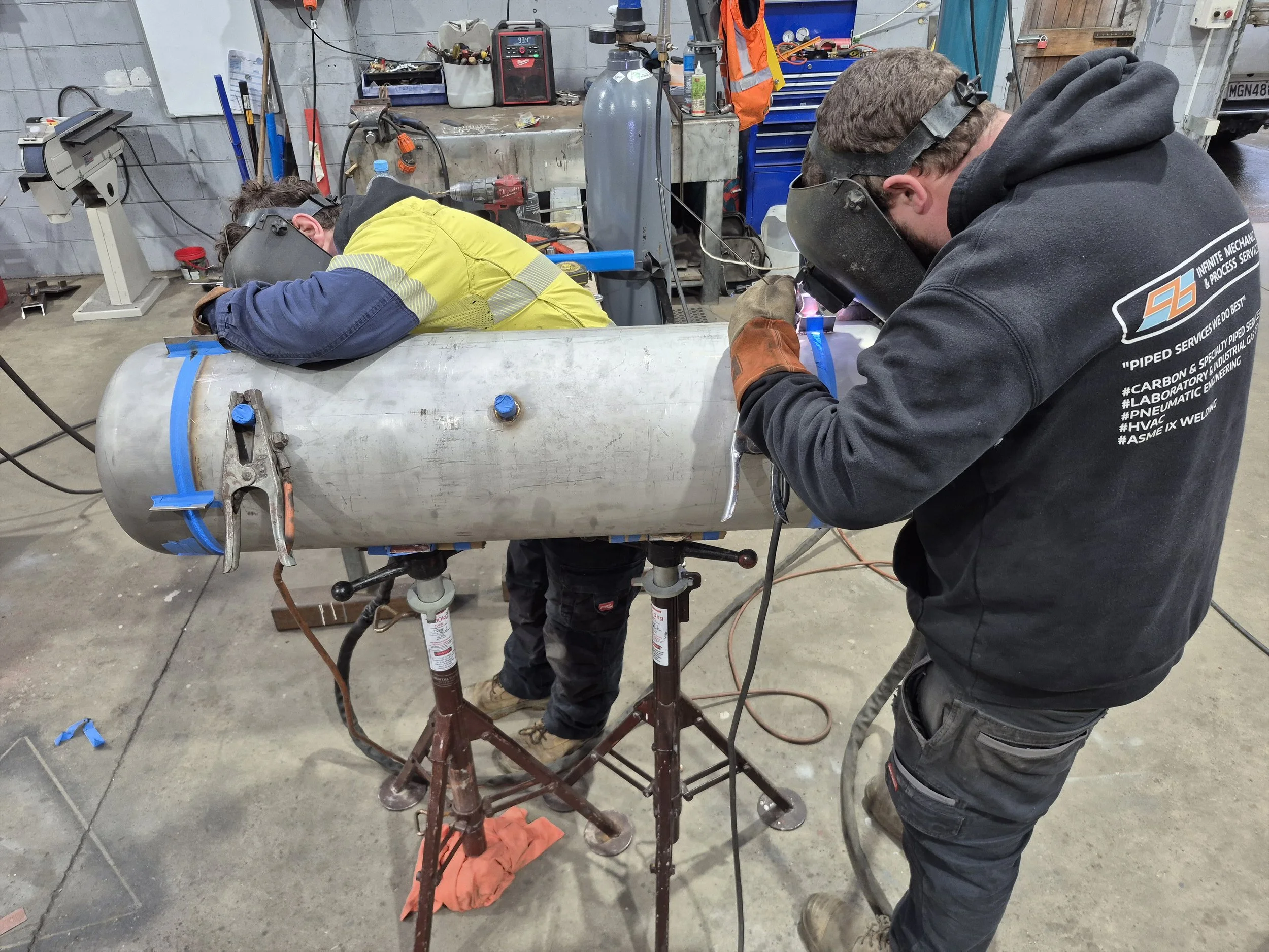 Two workers welding a large metal pipe in a workshop, both wearing safety goggles and protective gloves, with various tools and equipment in the background.