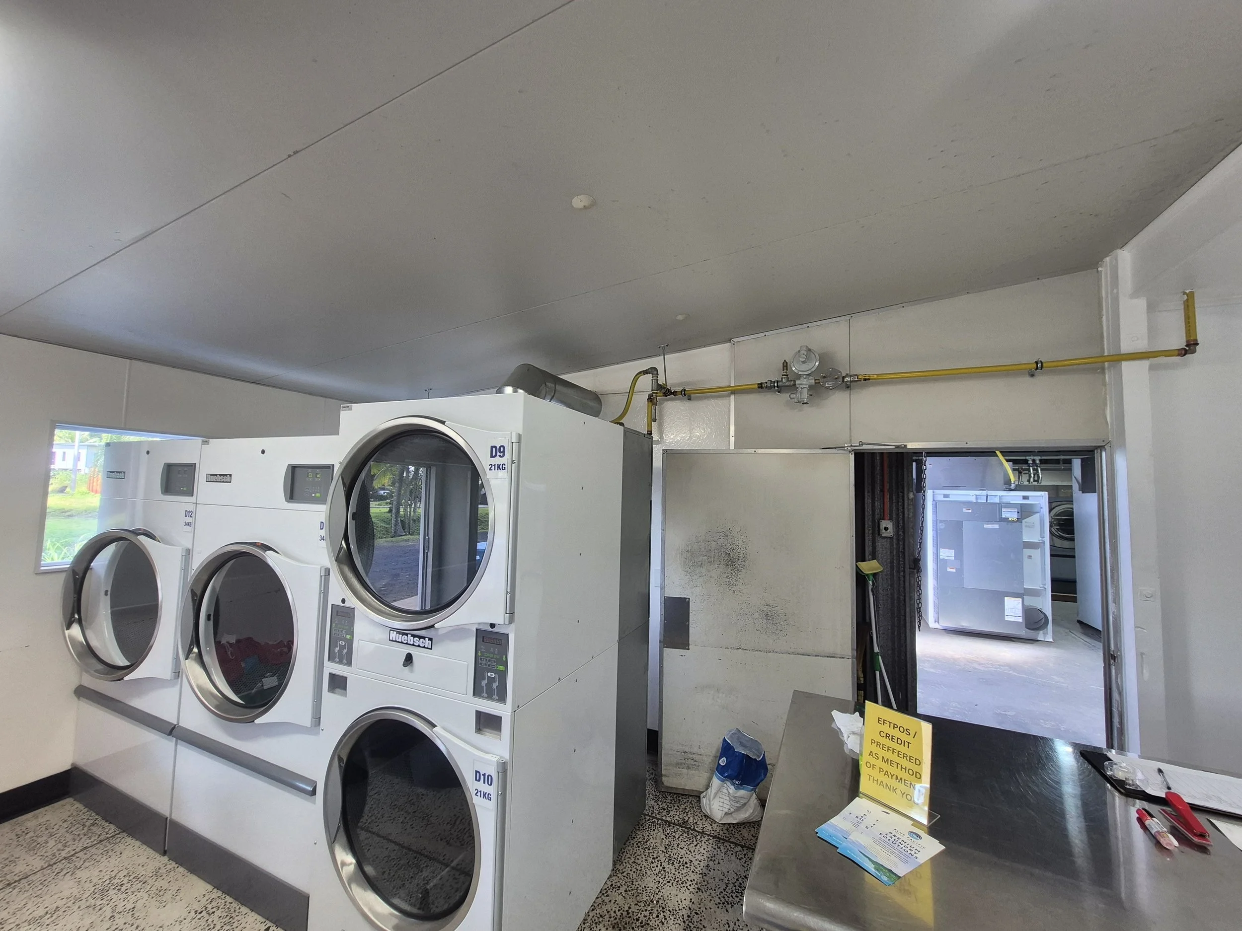 Coin-operated laundry area with three washing machines and a yellow notice on a metal counter, with cleaning supplies and a mop nearby.