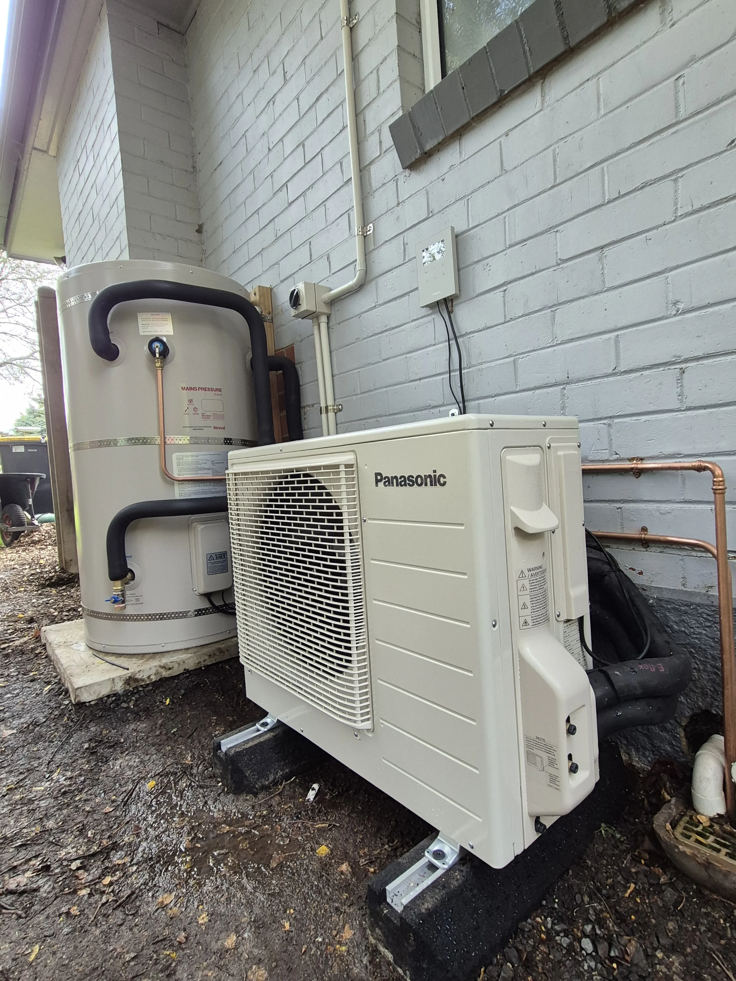 An outdoor air conditioning unit installed next to a wall with exposed pipes and electrical wiring, with a gray brick wall in the background.