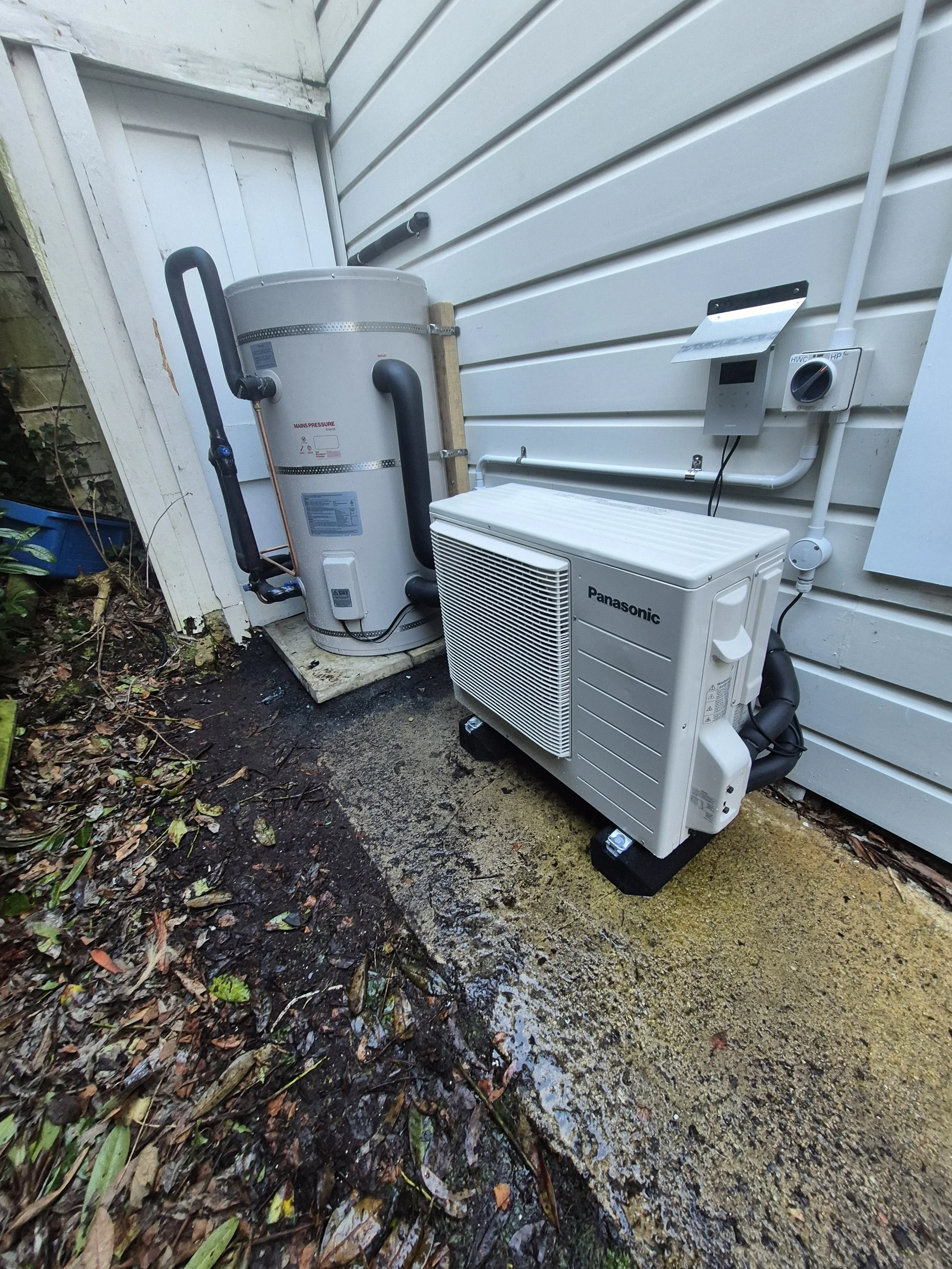 Outdoor air conditioning and water heater equipment installed against the side of a house, with pipes and control units visible, on a concrete slab surrounded by soil and fallen leaves.