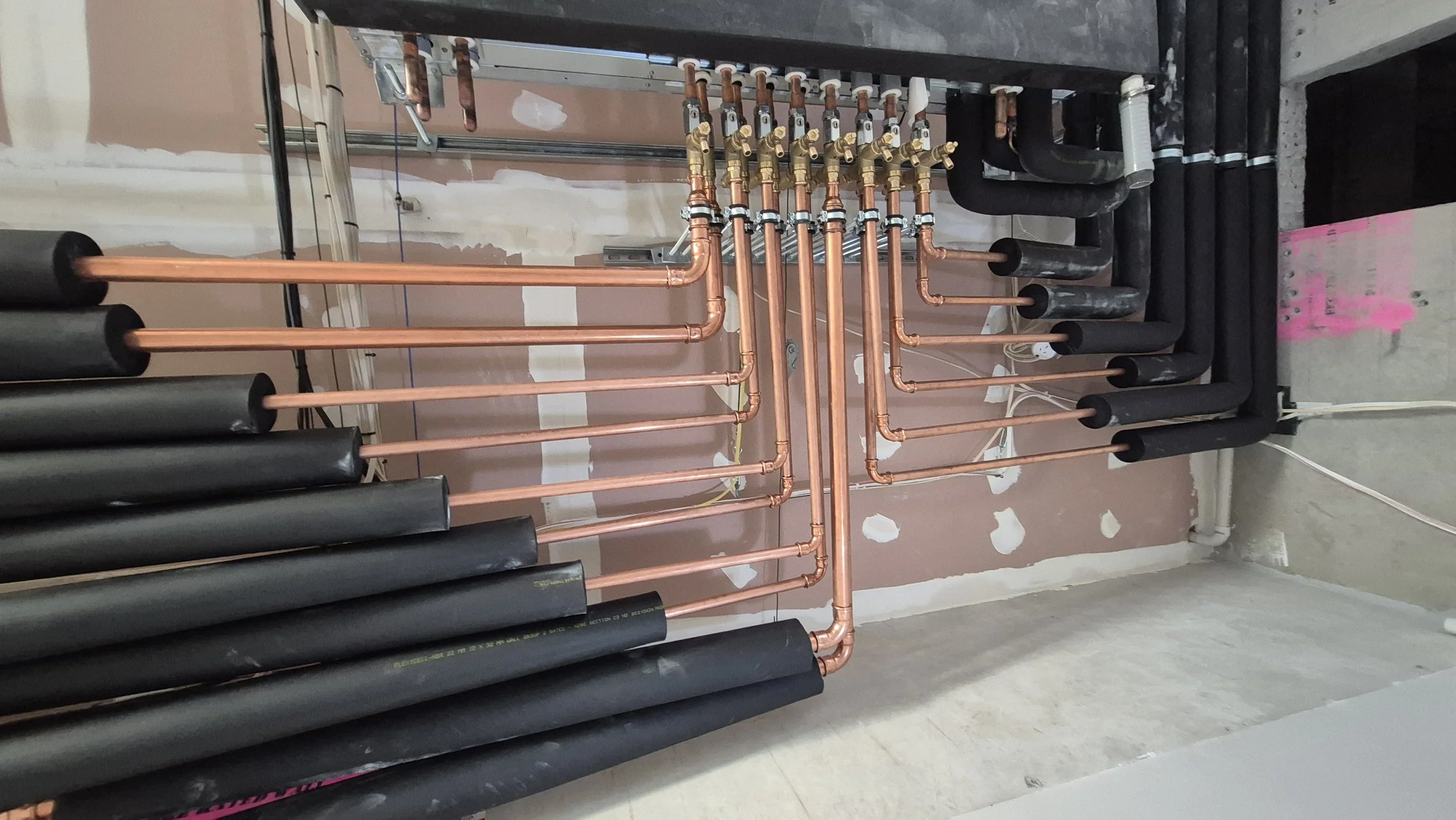 Copper plumbing pipes with black insulation and multiple valves installed on a wall in a building under construction or renovation.