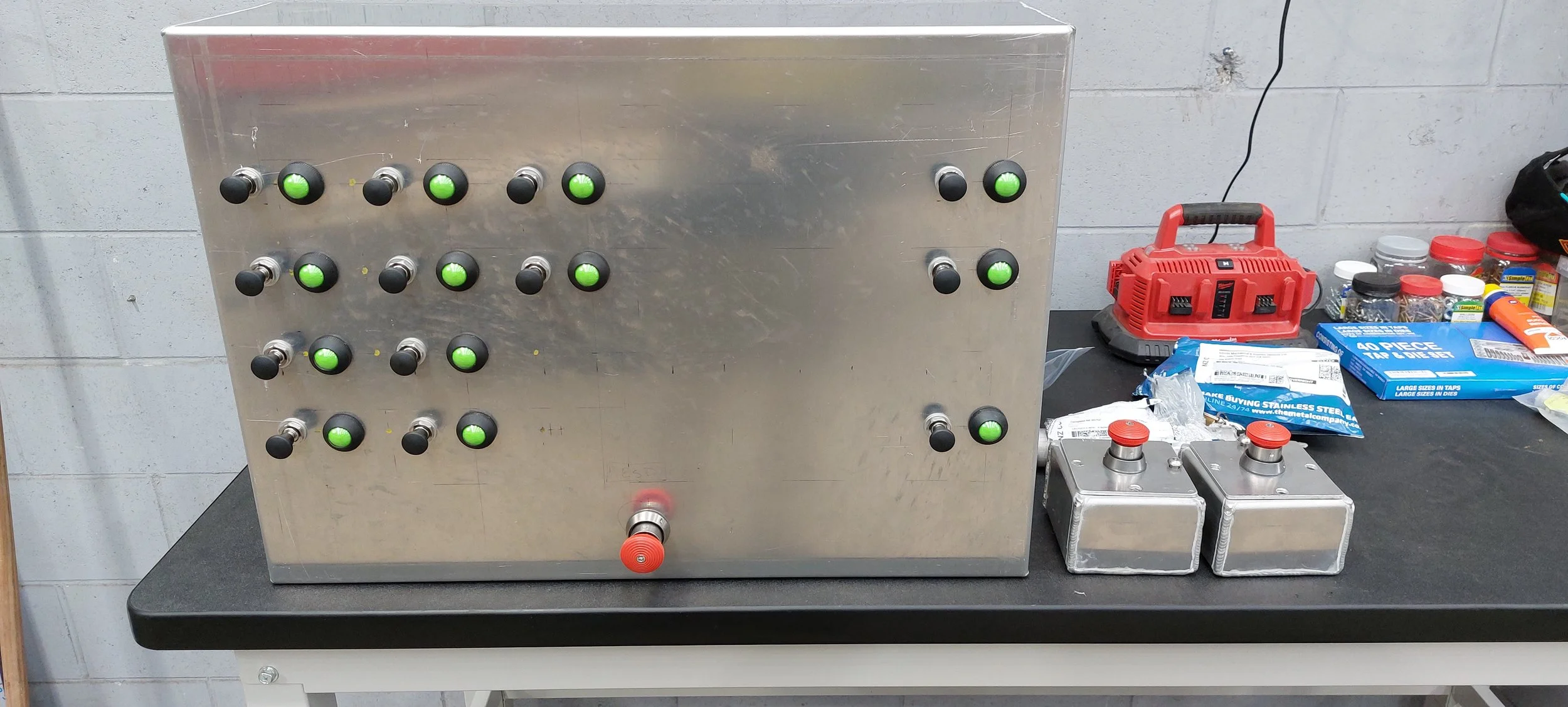 A stainless steel control box with multiple black and green push buttons on a black workbench. To the right, there is a red toolbox, small bottles, packets, and containers, along with metal boxes with red knobs. The background is a gray cinder block 