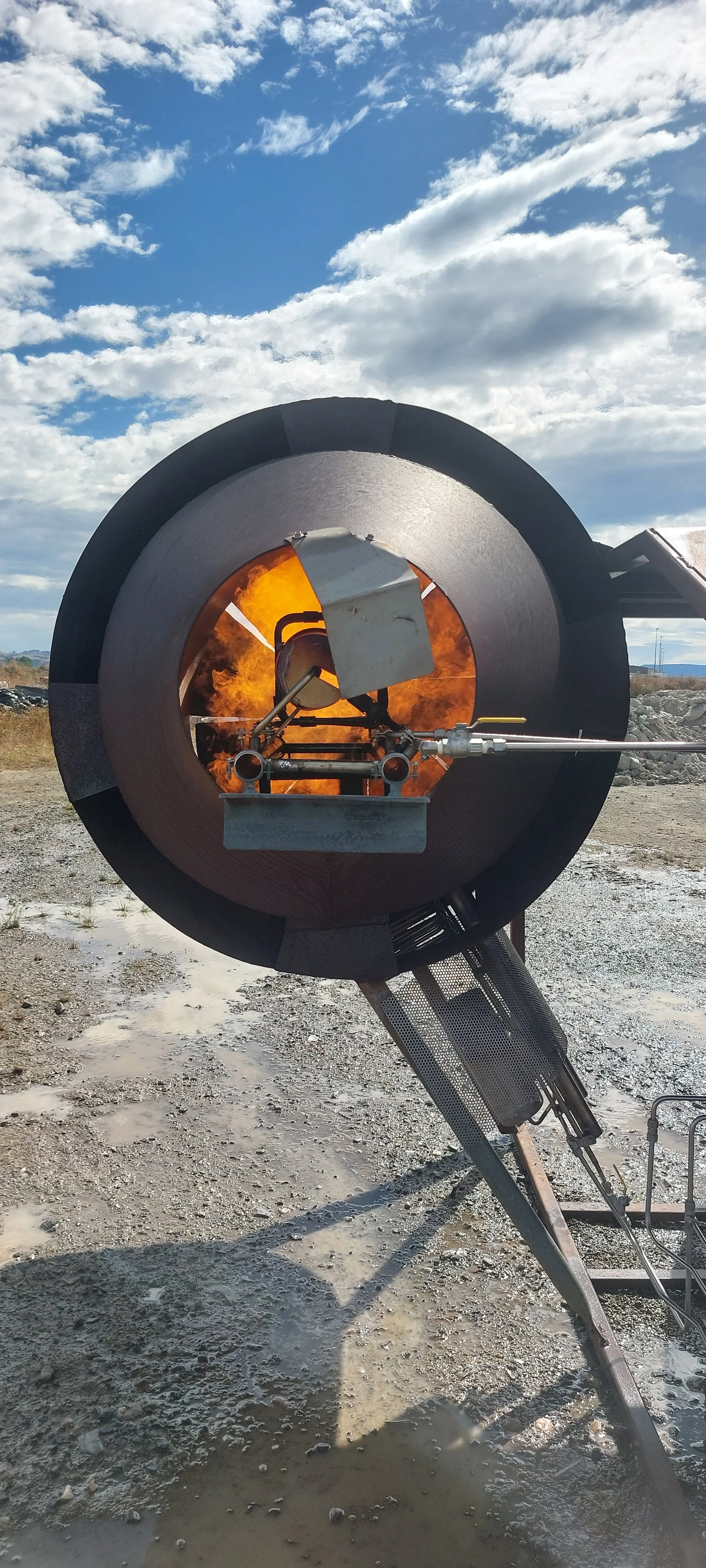 An outdoor scene with a large rocket engine with flames coming out from the nozzle, against a partly cloudy sky.