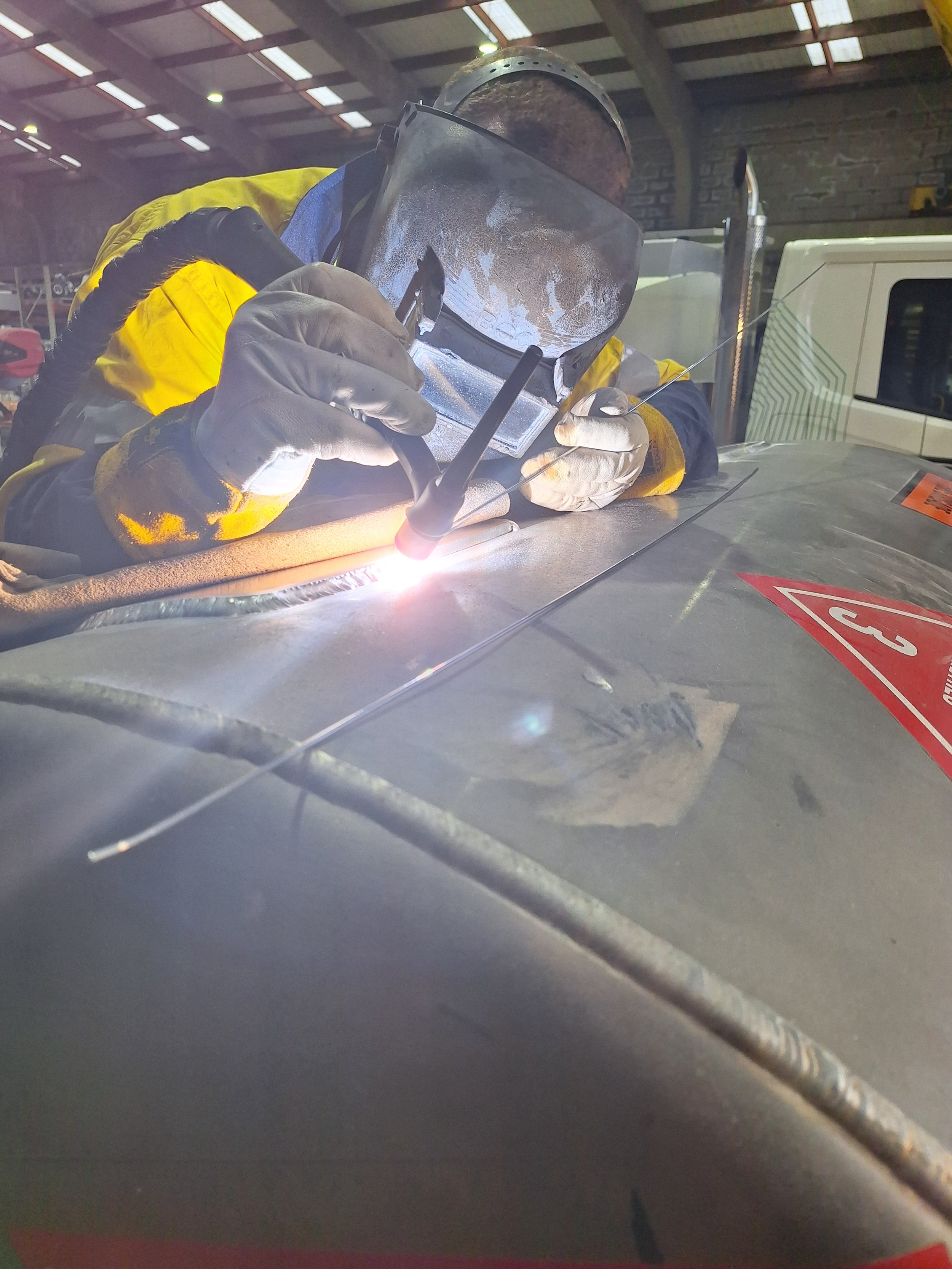 Worker welding metal on a vehicle in a workshop, wearing protective gear including a welding helmet and gloves.