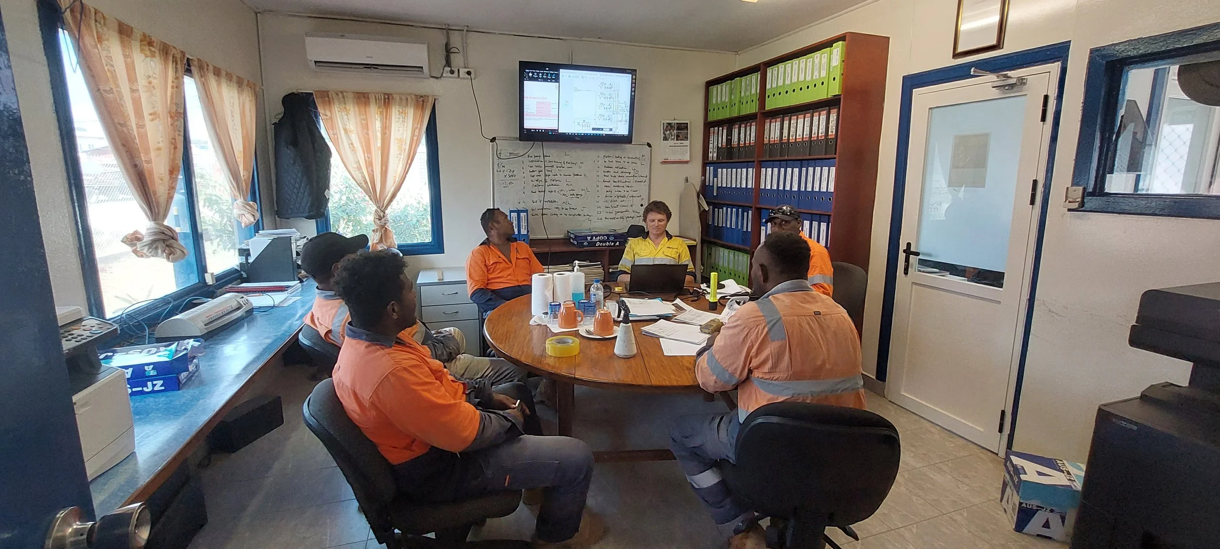 Six workers, wearing orange and yellow safety uniforms, seated around a conference table in a meeting room, with a whiteboard and a television screen showing a diagram behind them.
