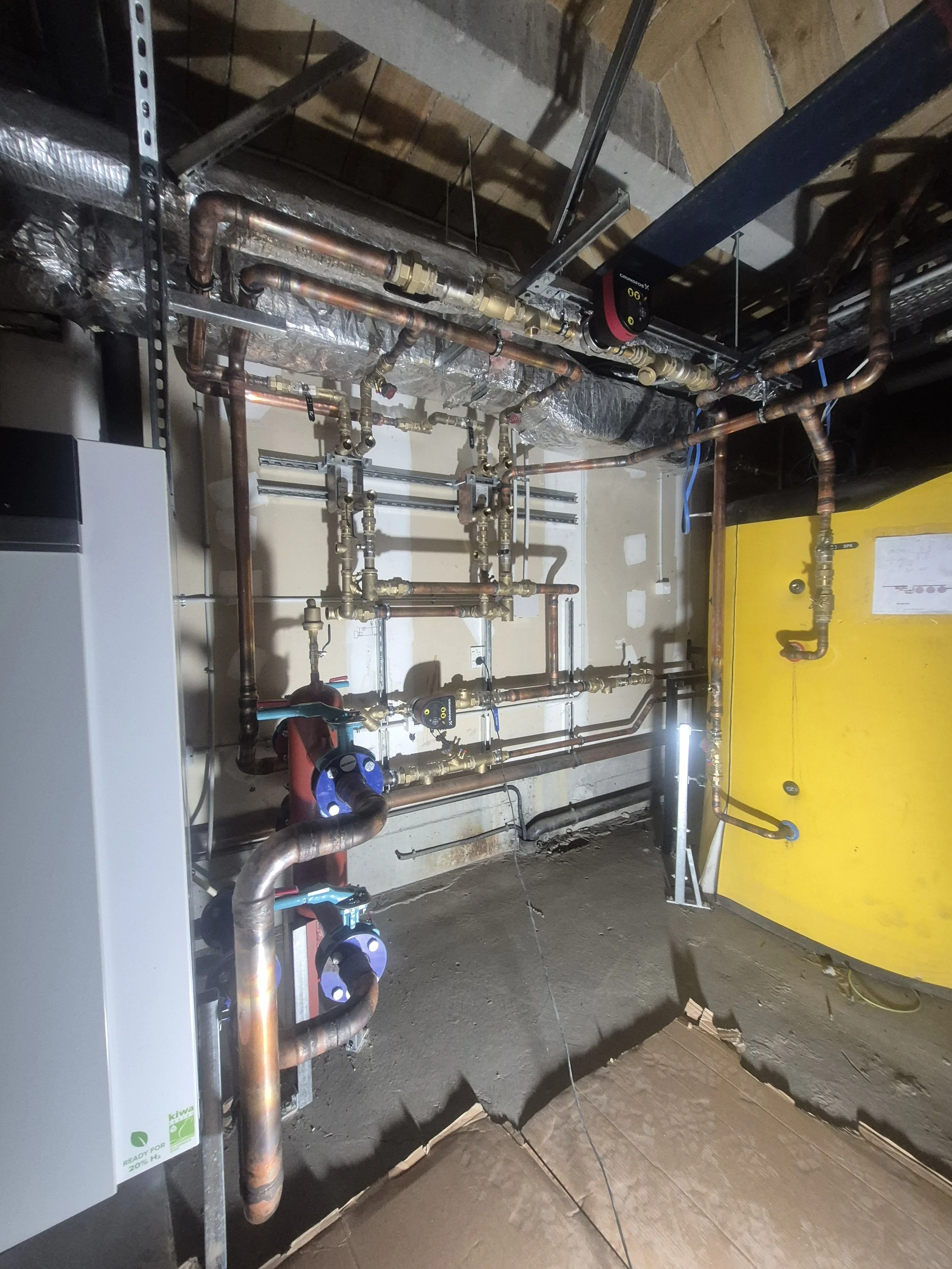 A complex network of copper and brass pipes, valves, and fitting fixtures installed on a wall in a building, with insulation and yellow equipment in the background, likely part of a plumbing or HVAC system under construction or maintenance.