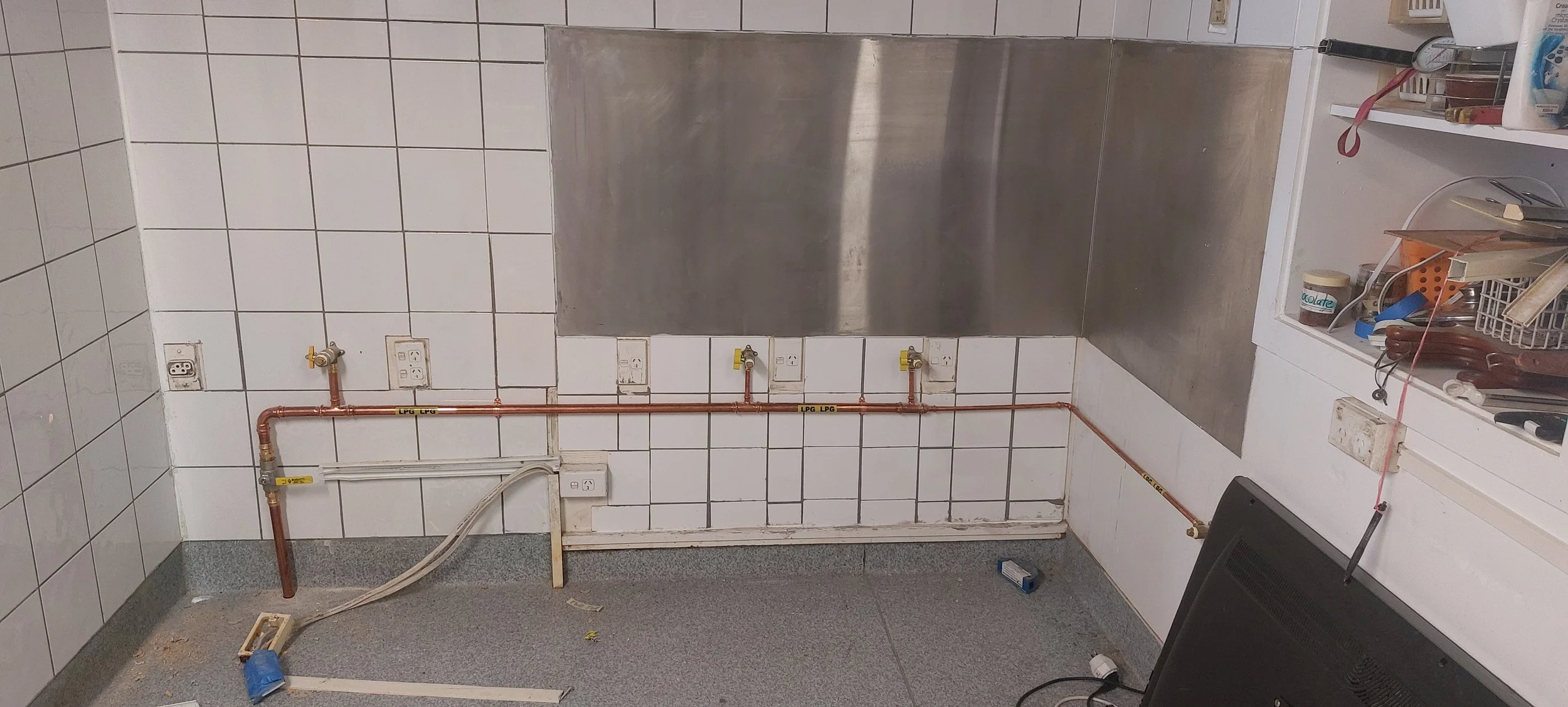 A kitchen with white tiled walls and a gray countertop. There are three LPG gas connections with copper pipes running along the wall. To the right, there are kitchen shelves with various items. A black electronic device is on the floor in the corner.