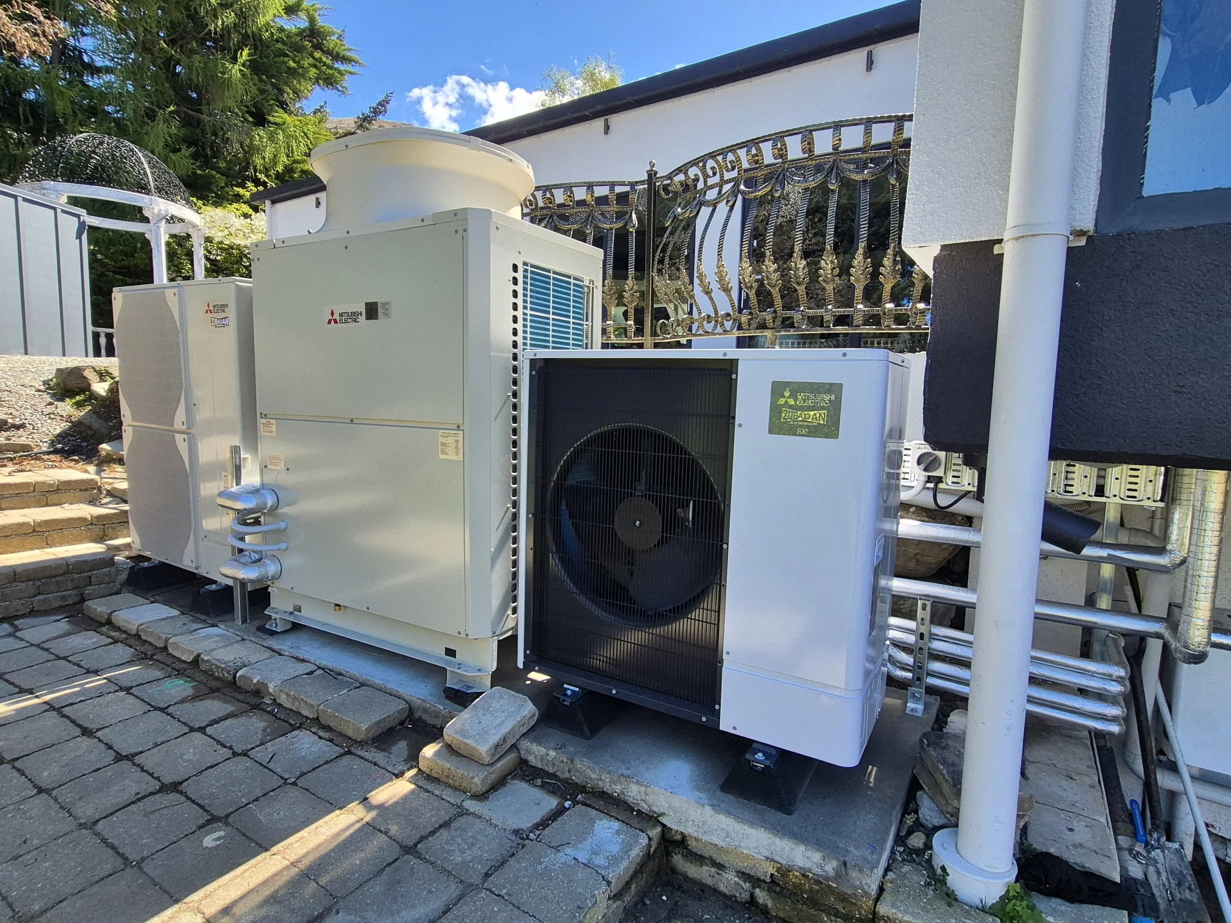 Outdoor installation of multiple HVAC units, including Mitsubishi Electric and Zubadan brands, placed on a paved surface next to a house with a white exterior and decorative metal railing. The scene shows a clear sky with some clouds.