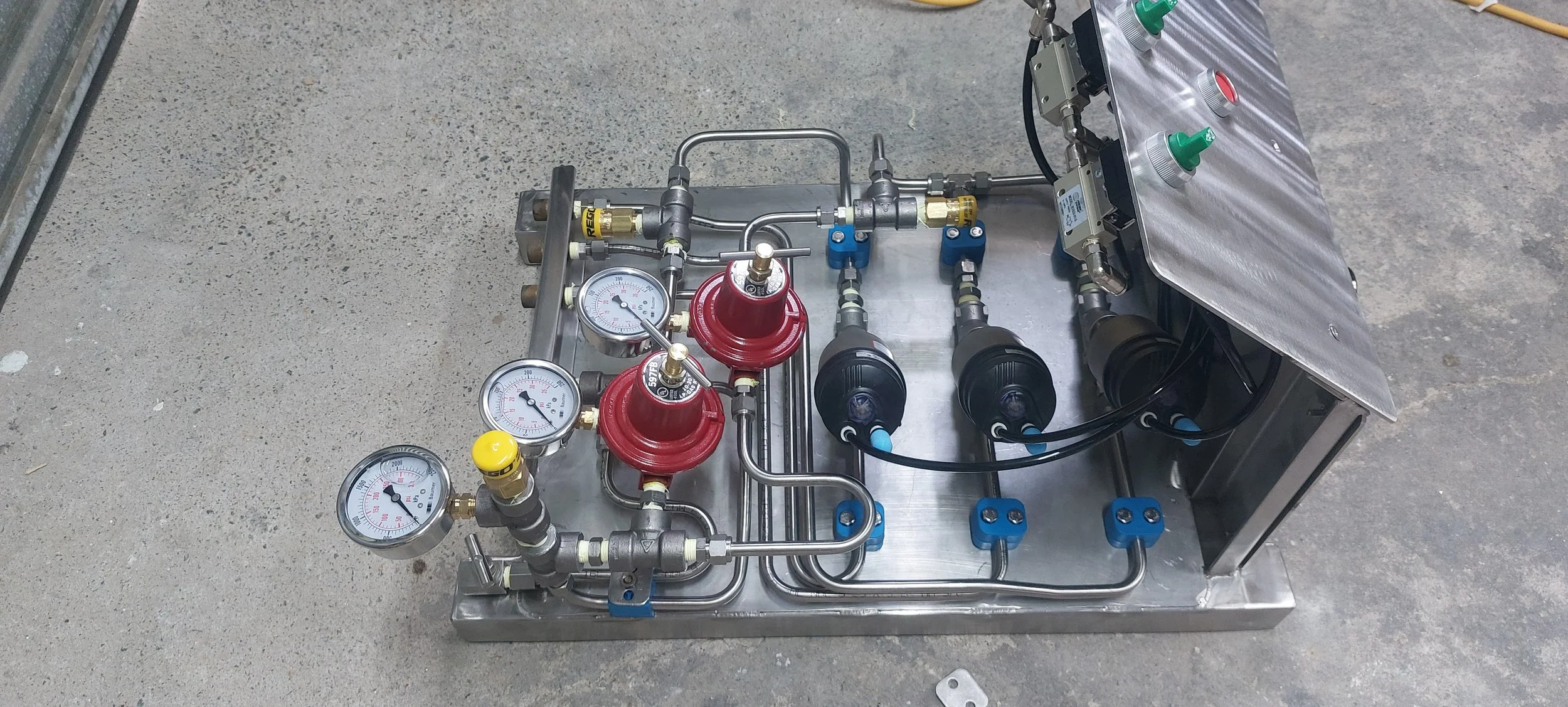 Industrial machinery with gauges, tubes, valves, and switches on a metal base, set on a concrete floor.