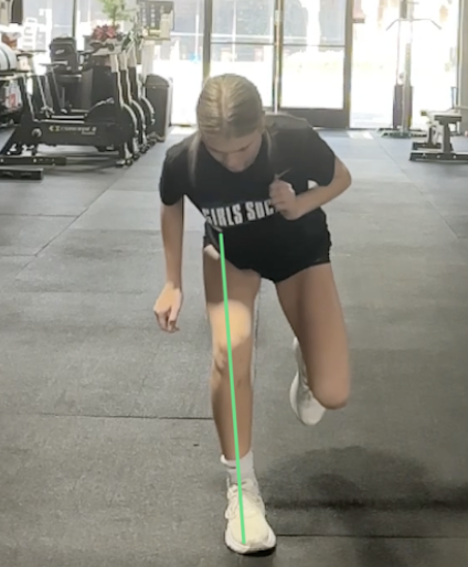 A woman in a gym performing a lunge exercise on a gray mat, with gym equipment in the background.