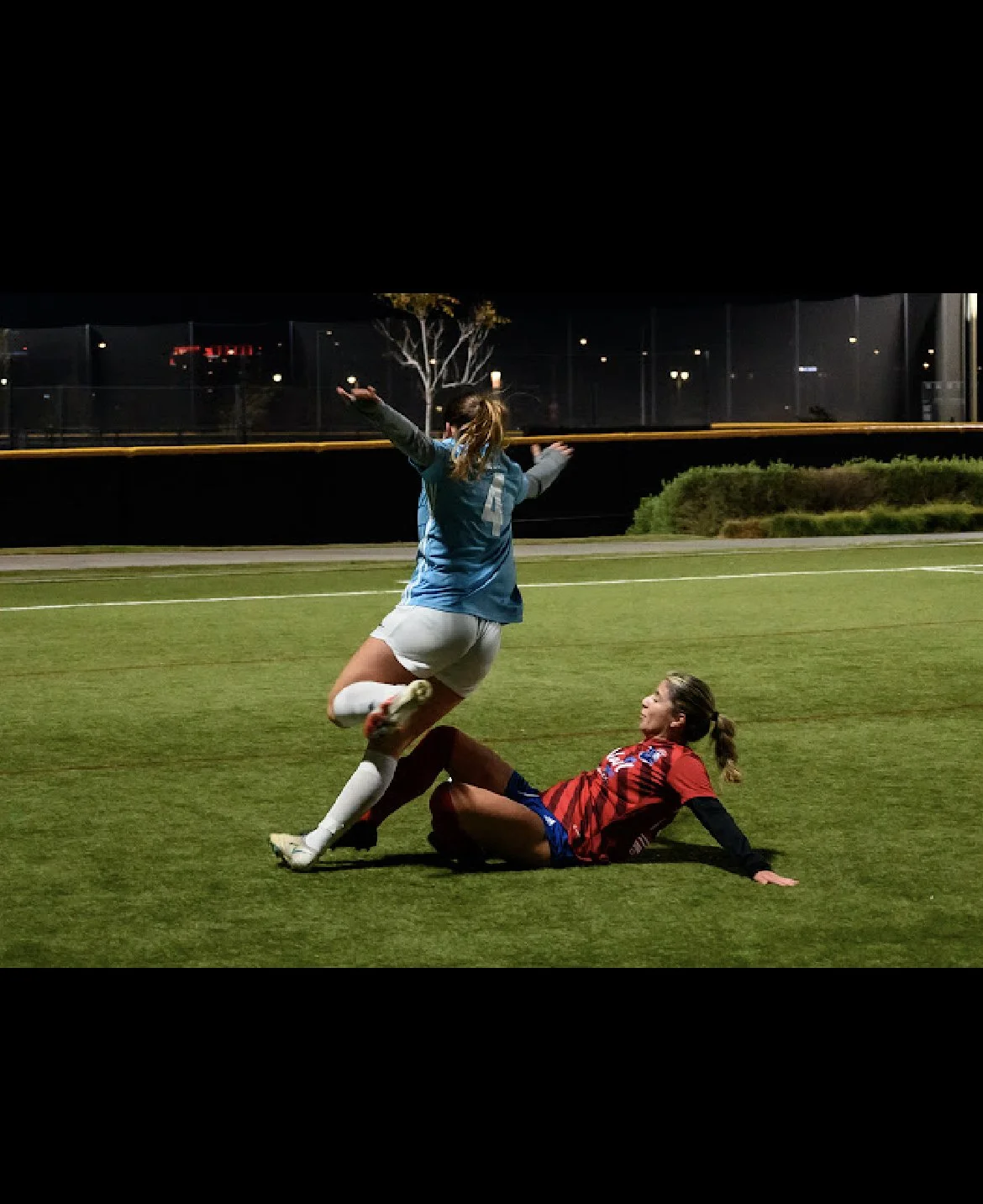 Two female soccer players on a field at night, one in a blue jersey and white shorts jumping over the other in a red and black jersey, who is sitting on the ground. The player in red is looking up at the player in blue.