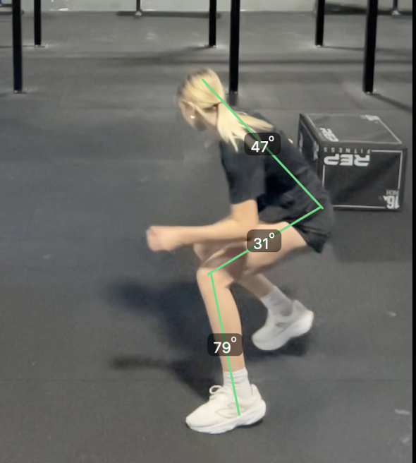 A woman in an athletic shirt and shorts is in a squatting position in a gym.