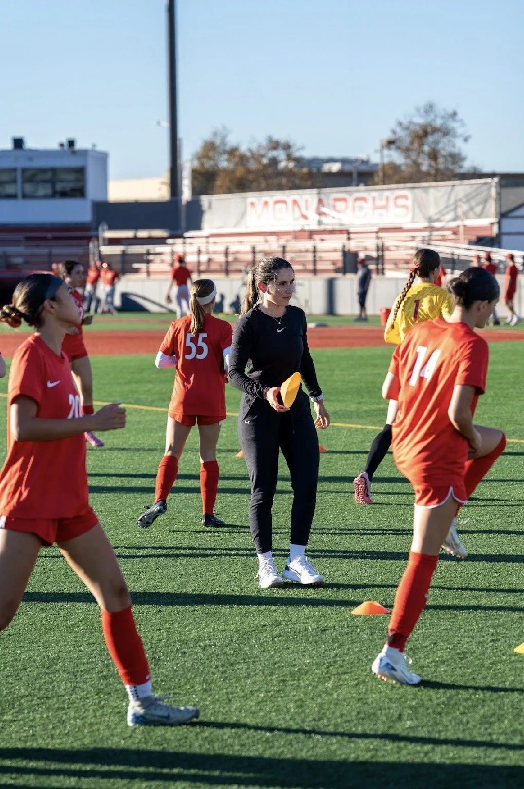 Women soccer players warming up on the field with a coach holding a yellow training cone during daytime, in a stadium with a sign that says 'MONARCHS' in the background.