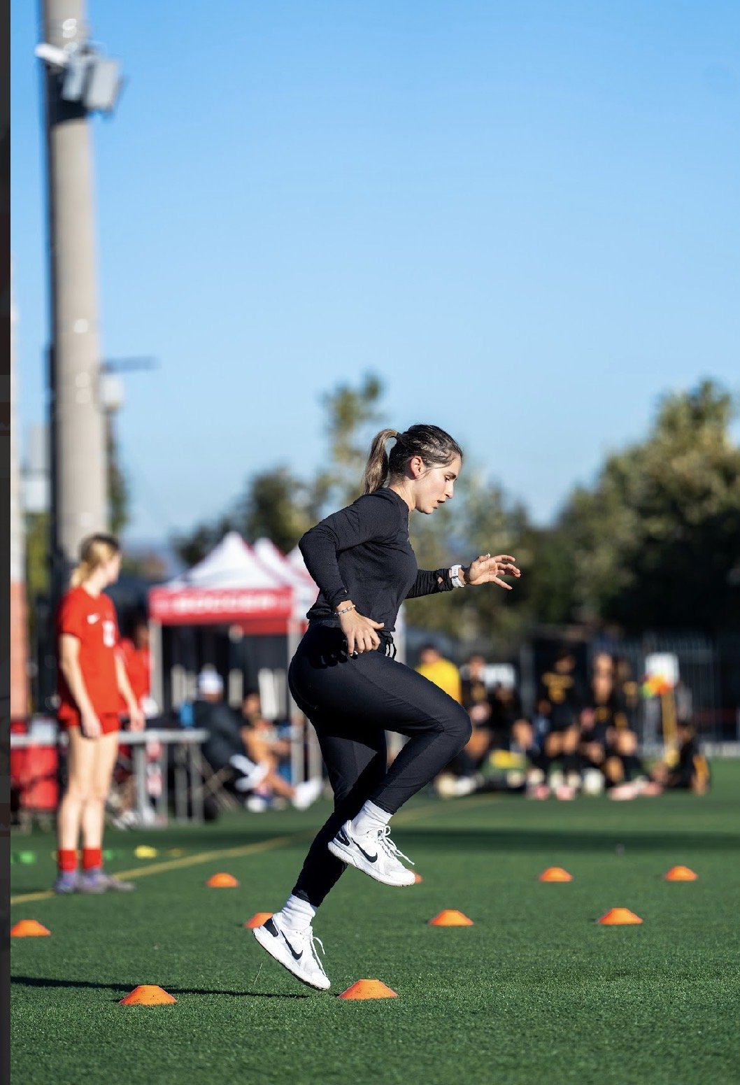 A woman with a ponytail jumping over orange cones during an outdoor athletic training session, with a team and tents in the background.