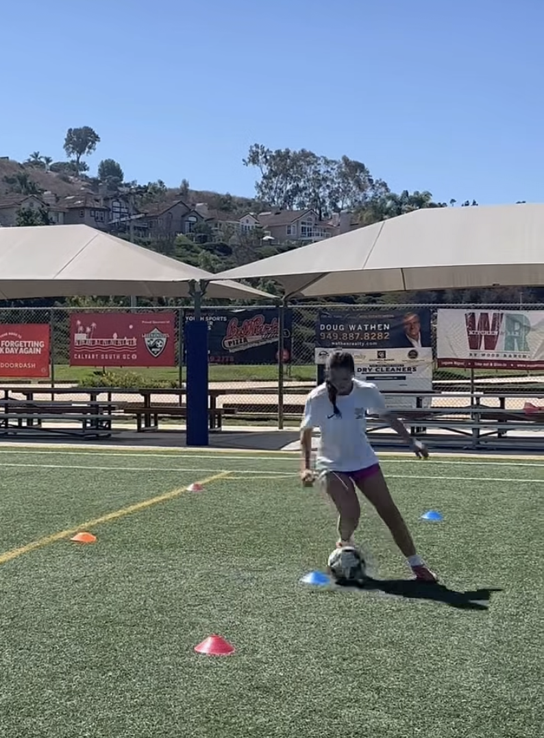 A girl practicing soccer on a field with orange and blue cones, wearing a white shirt and pink shorts, with a soccer ball at her feet.