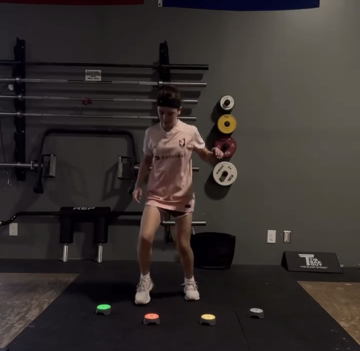 A woman in pink athletic clothing in a gym, performing an exercise on a black mat with colored markers, with weightlifting equipment and bumper plates in the background.