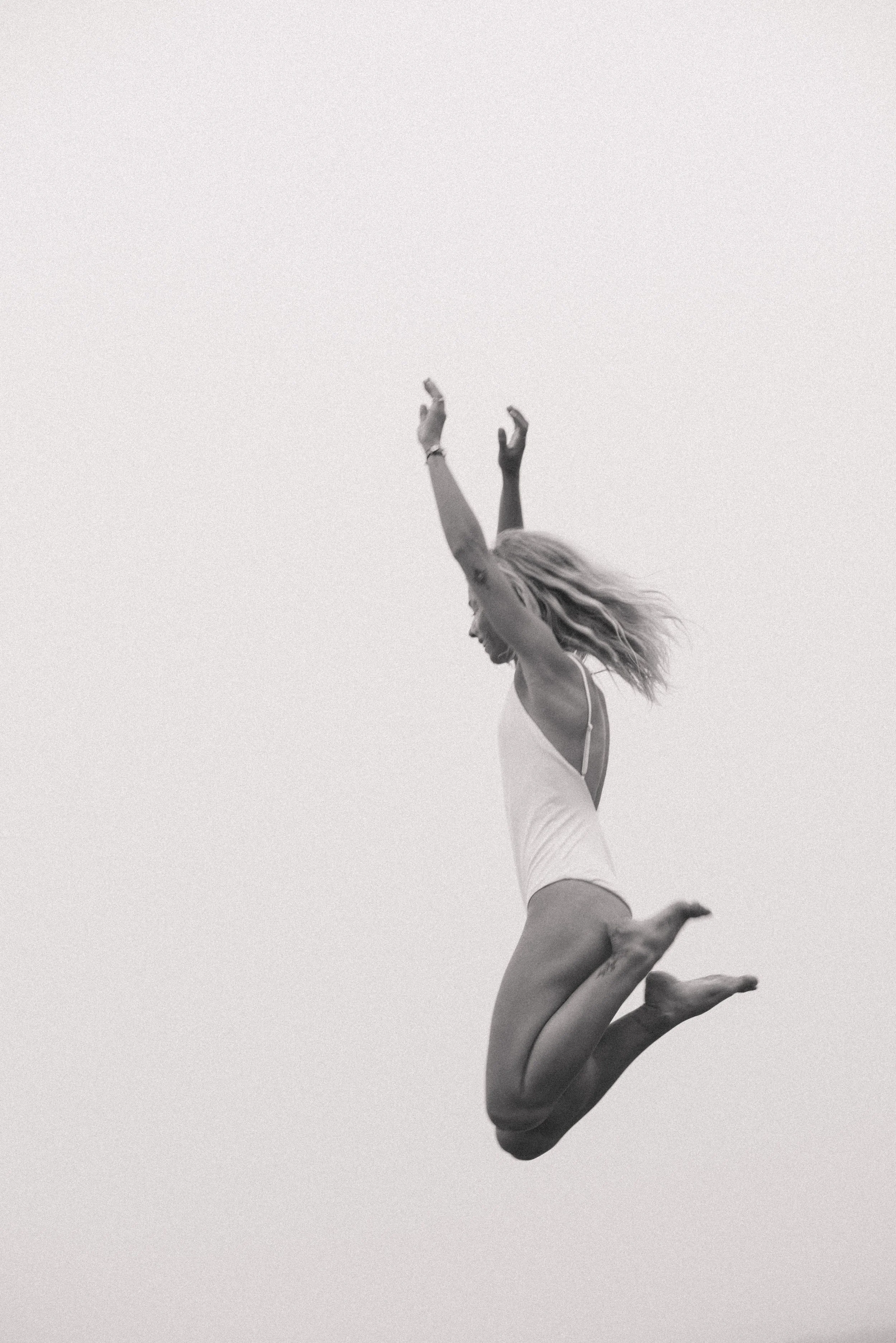 A woman in a white swimsuit is captured mid-air jump against a plain, light background with her arms raised and hair flowing.