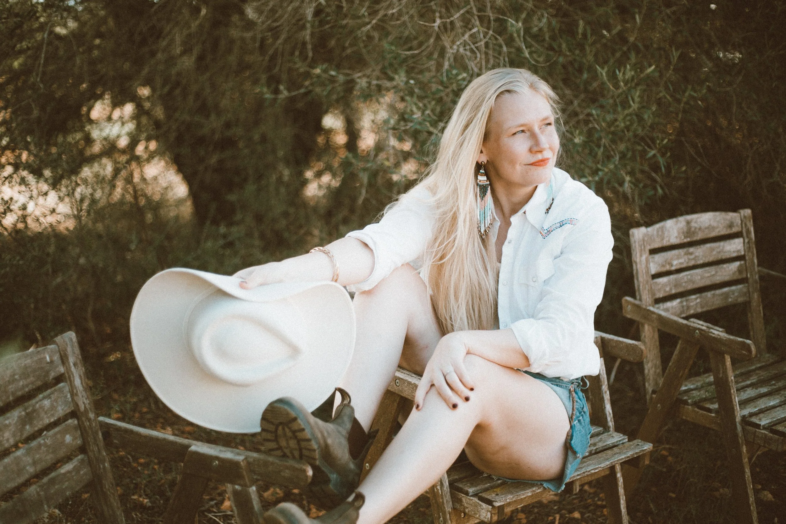 A woman with long blonde hair sitting on a wooden chair outdoors, holding a white cowboy hat, wearing denim shorts and a white shirt, with trees in the background.