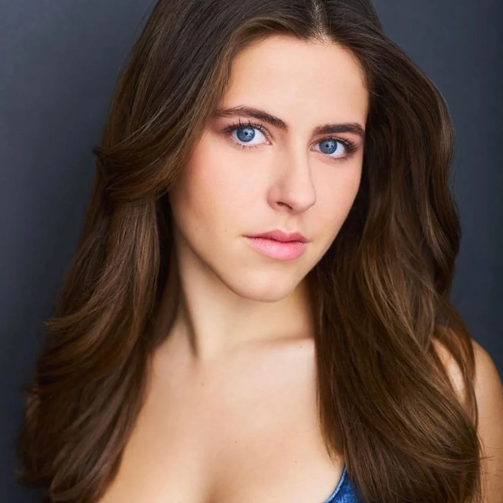 Close-up portrait of a young woman with long, wavy brown hair and blue eyes, against a dark background.