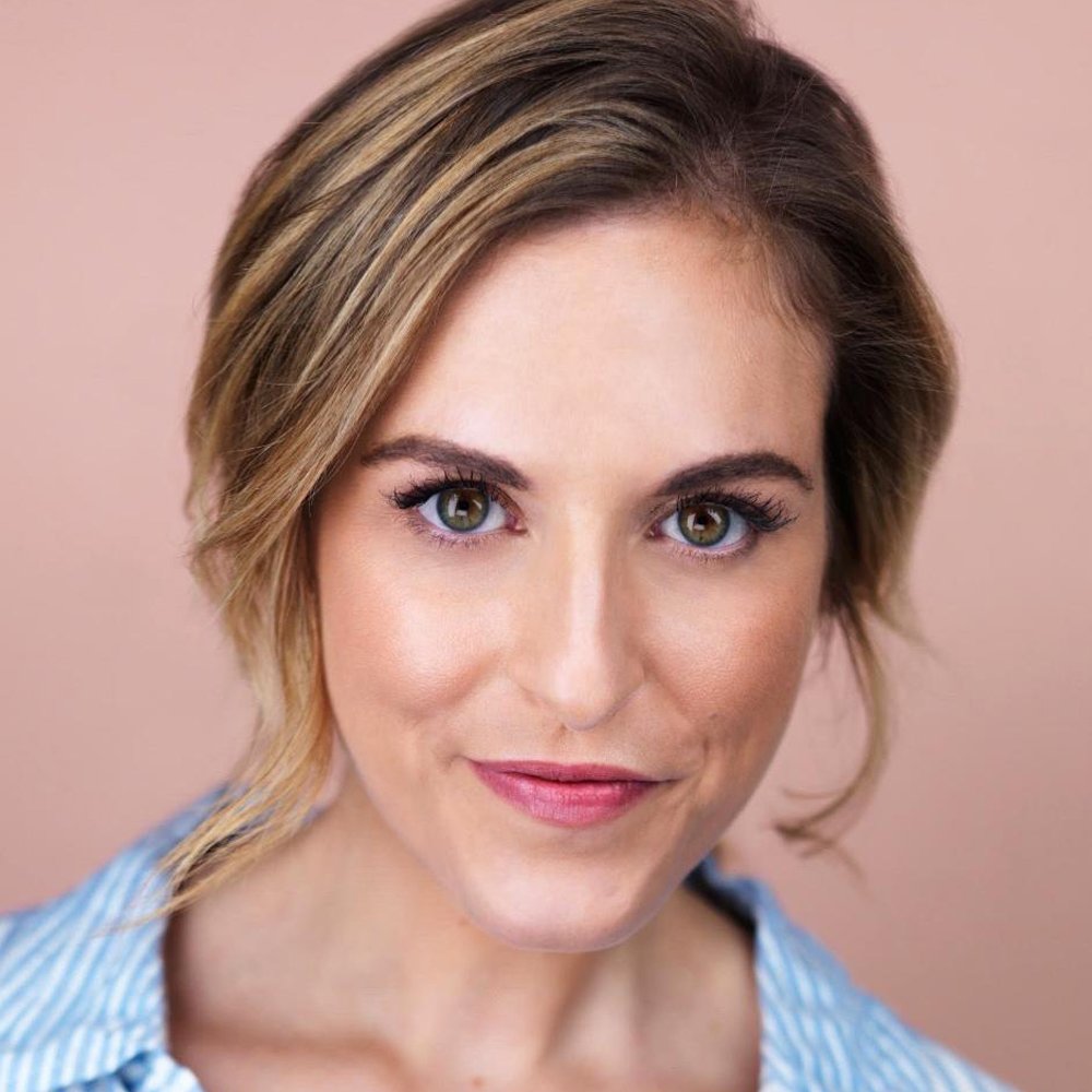 Close-up portrait of a woman with short light brown hair and hazel eyes, wearing a light blue and white striped shirt, against a soft pink background.