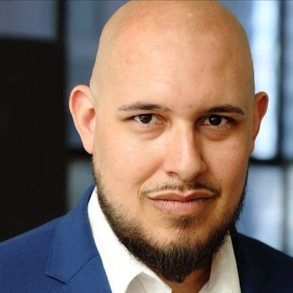 Close-up of a man with a bald head, beard, and mustache, wearing a dark blue suit and white shirt, looking into the camera with a neutral expression.