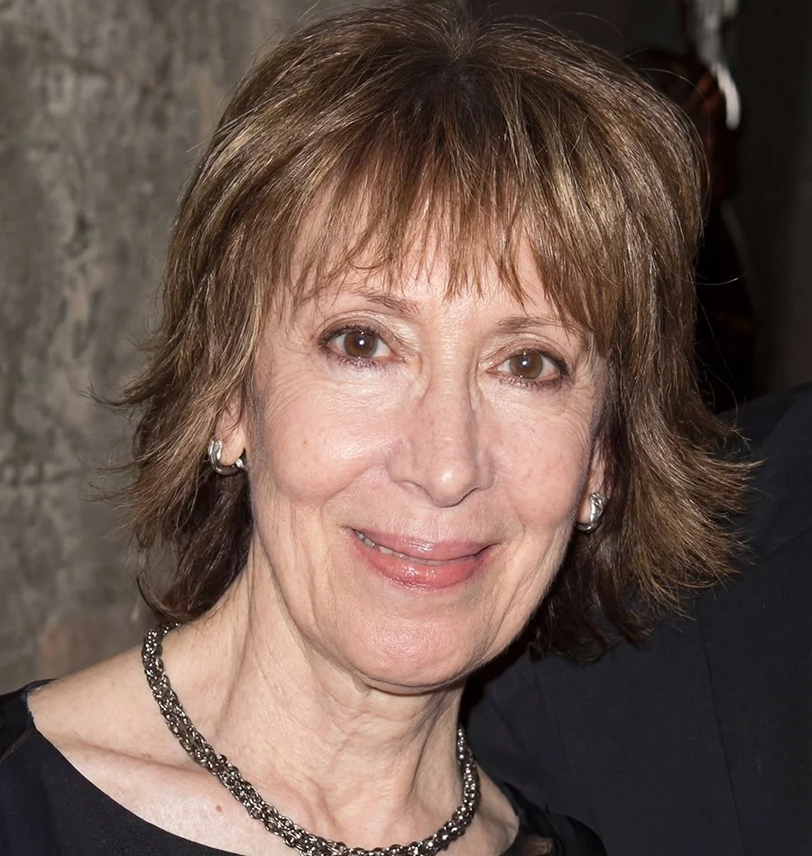 Close-up of a middle-aged woman with short, layered brown hair, wearing earrings, a necklace, and a black top, smiling in front of a stone wall.