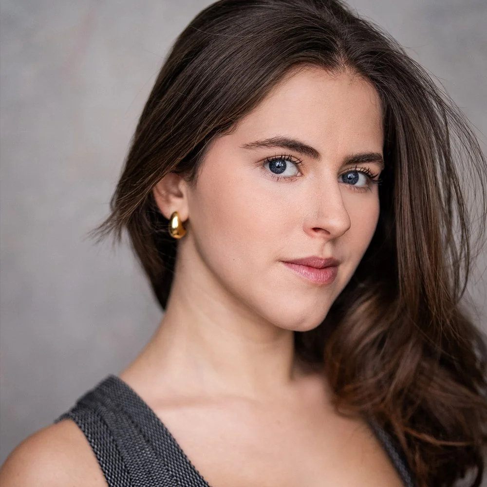 Close-up portrait of a young woman with long brown hair, blue eyes, wearing gold earrings and a sleeveless gray top, looking at the camera.