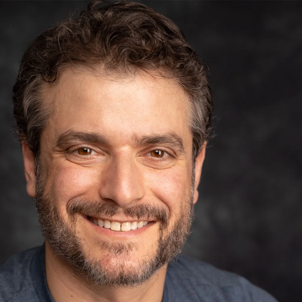 Close-up portrait of a smiling man with brown eyes, curly dark hair, and a beard.