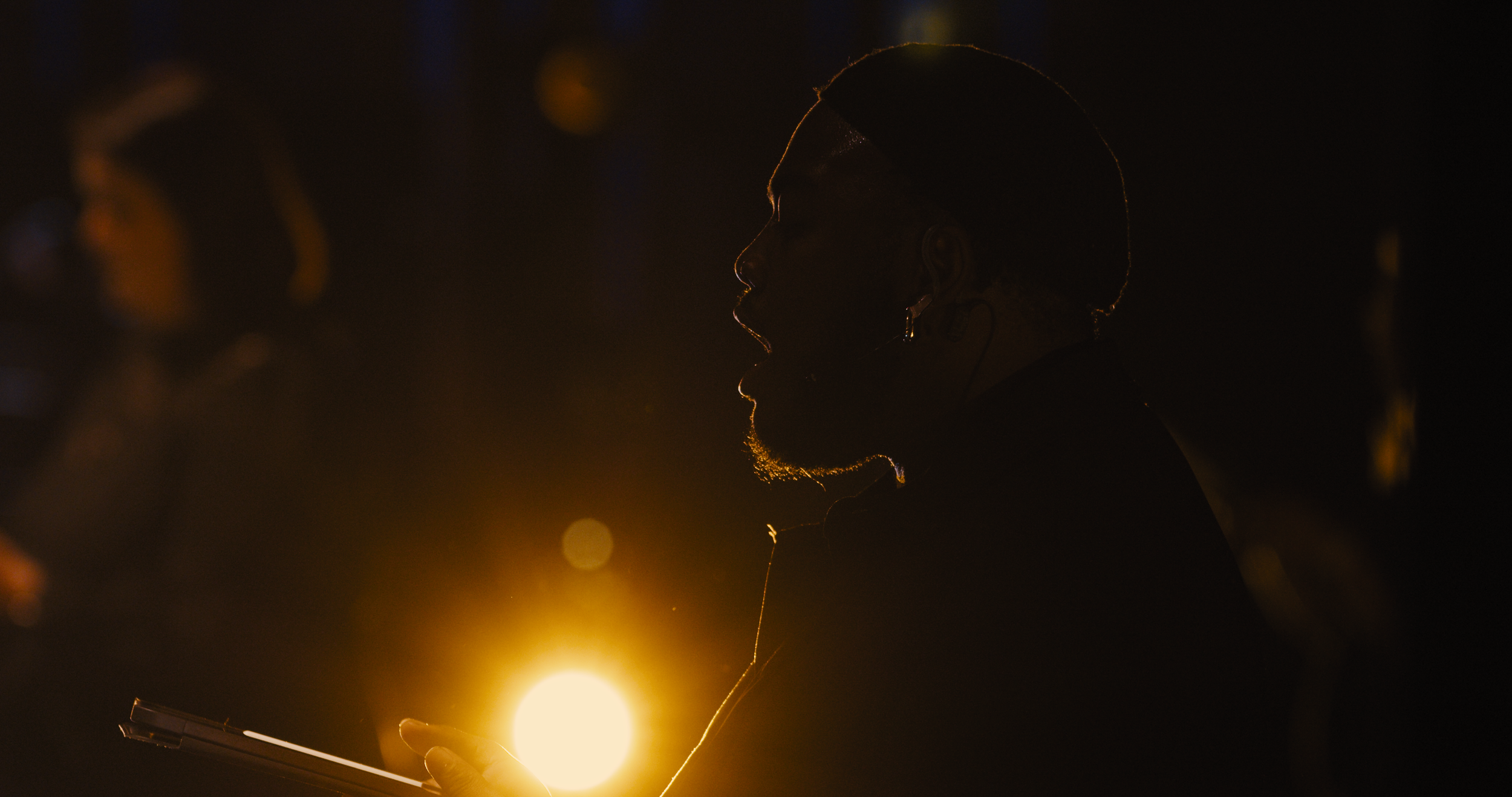 Silhouette of a person with a ponytail, looking at a device, backlit by a bright light, in a dark environment.