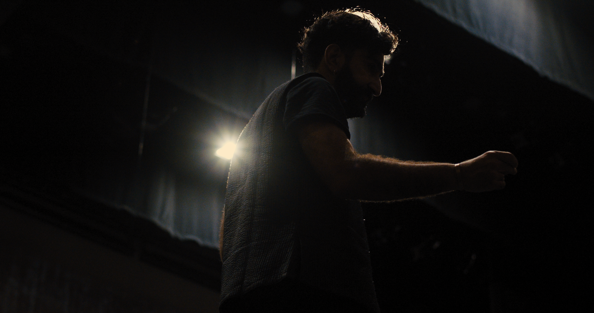 A man with a beard and glasses silhouetted against a bright light, with his hand extended forward, in a dark indoor setting.
