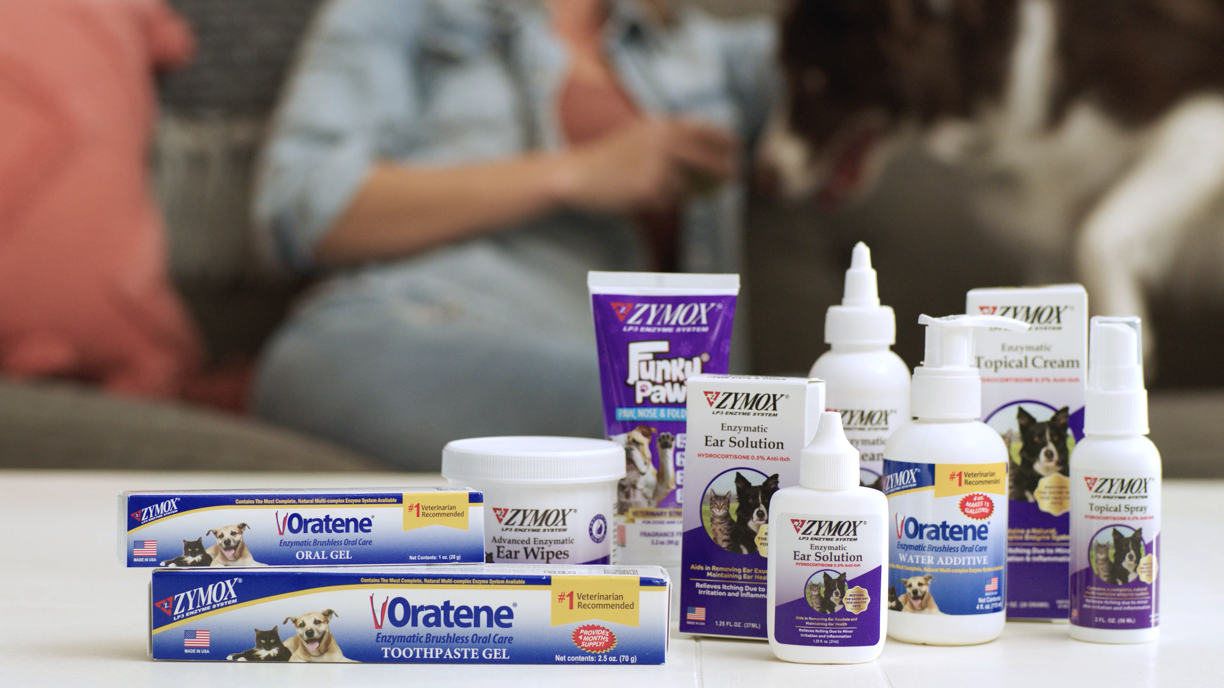 Various Zymox veterinary pet healthcare products, including oral gel, toothpaste, ear wipes, ear solution, topical cream, water additive, ear spray, and Funku Paw for dogs and cats, arranged on a table, with a person and a dog in the blurred backgrou