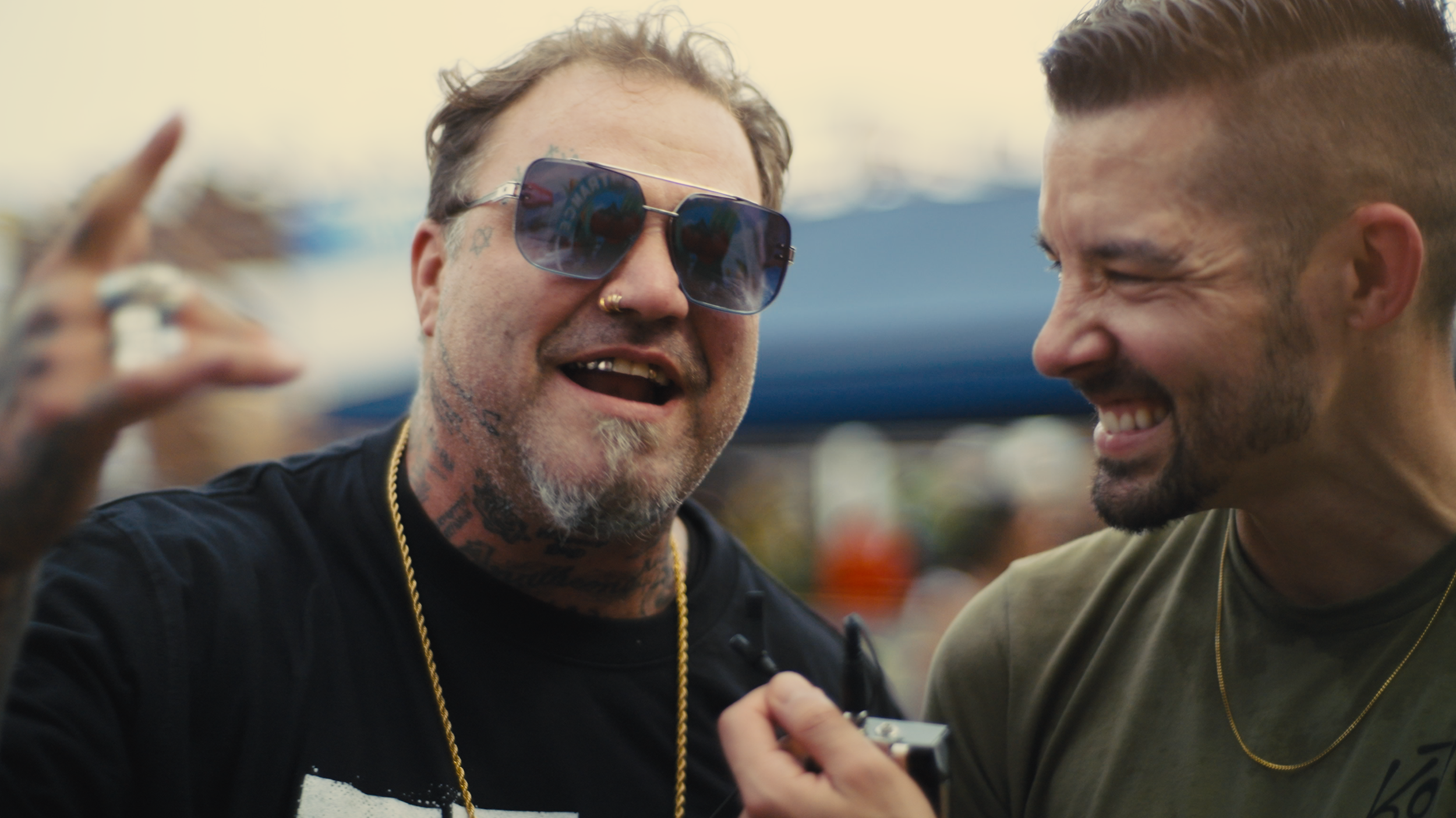 Two men are smiling and talking to each other, one holding a recording device. The man on the left is wearing sunglasses, has tattoos on his neck and face, and is dressed in black. The man on the right has short hair, a beard, and is wearing a green 