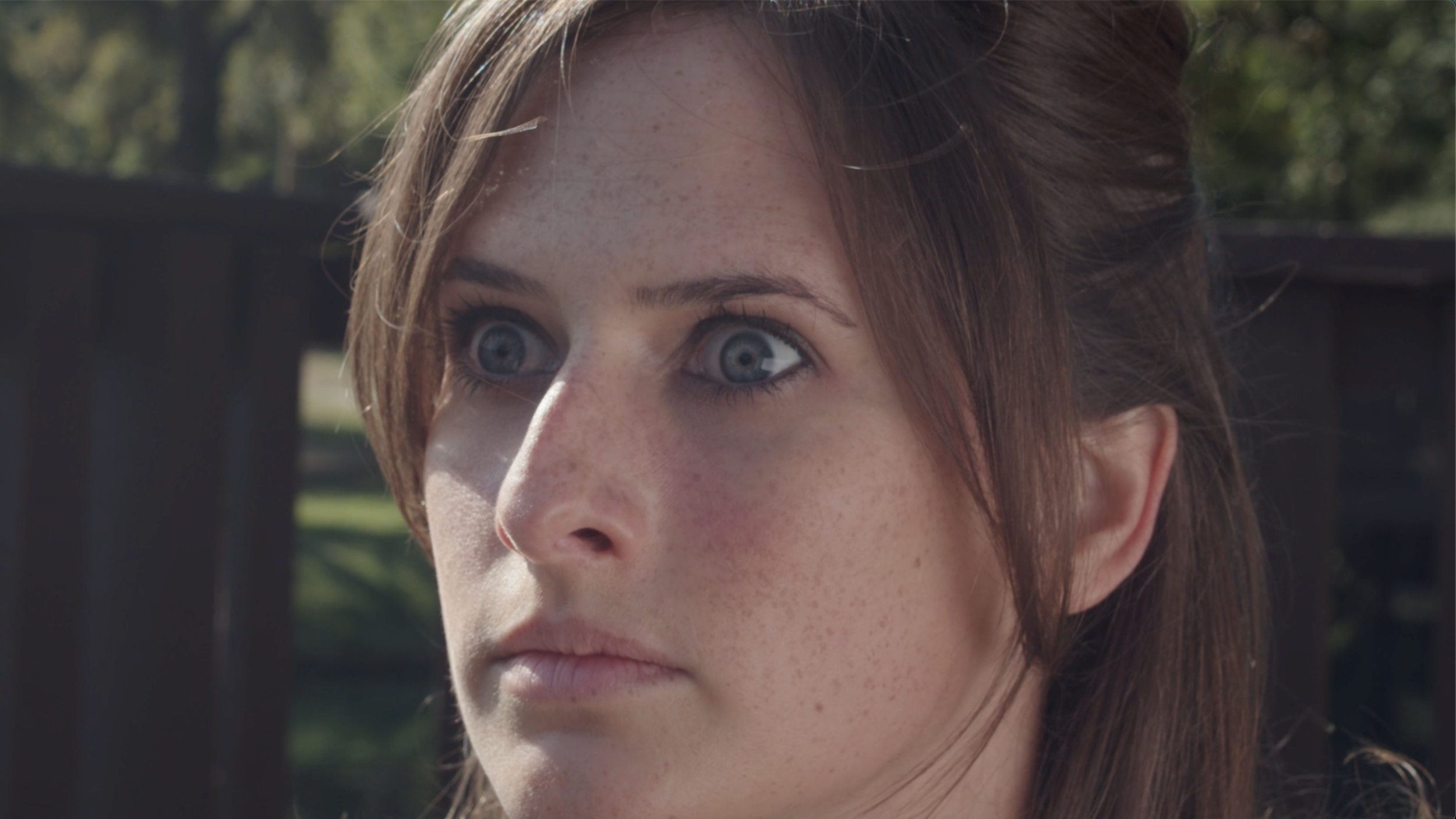 Close-up of a woman with blue eyes, freckles, and brown hair outdoors with a blurred fence and trees in the background.