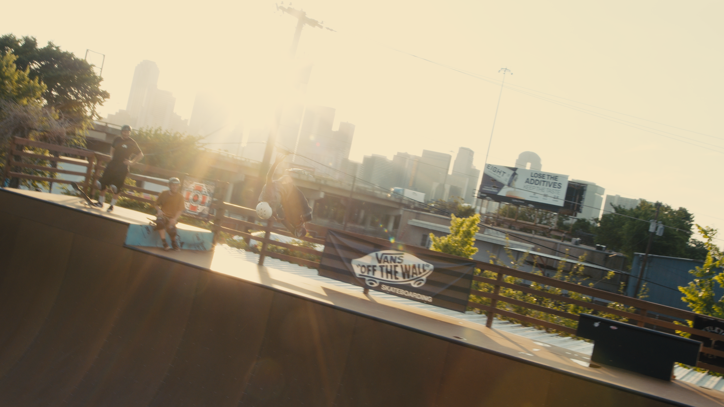 Skateboarders at an outdoor skate park with a city skyline in the background, posters, and banners, including a Vans 'Off the Wall' sign, during golden hour sunlight.