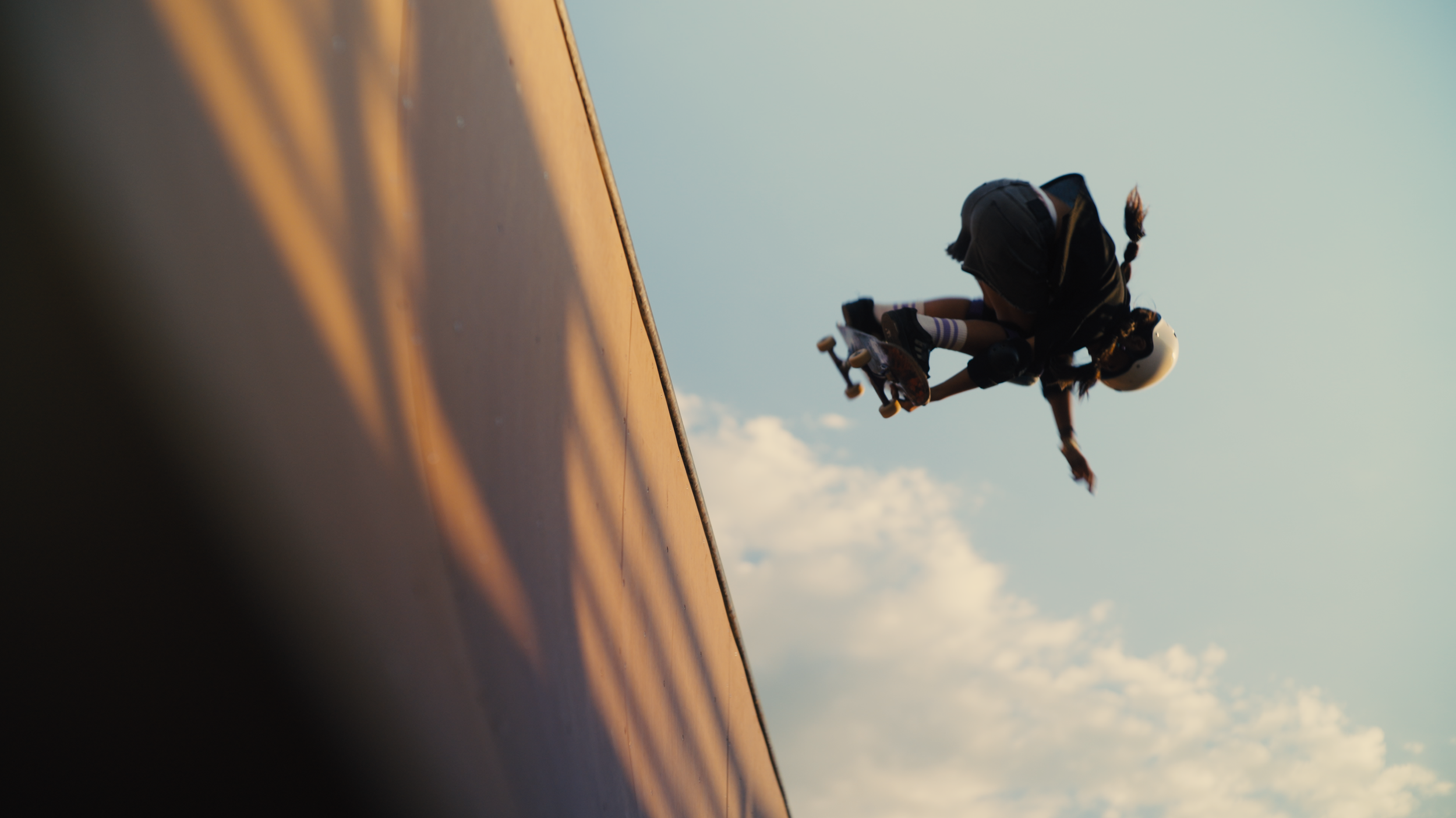A girl in a helmet performing a skateboarding trick at a skate park during sunset with a clear sky in the background.