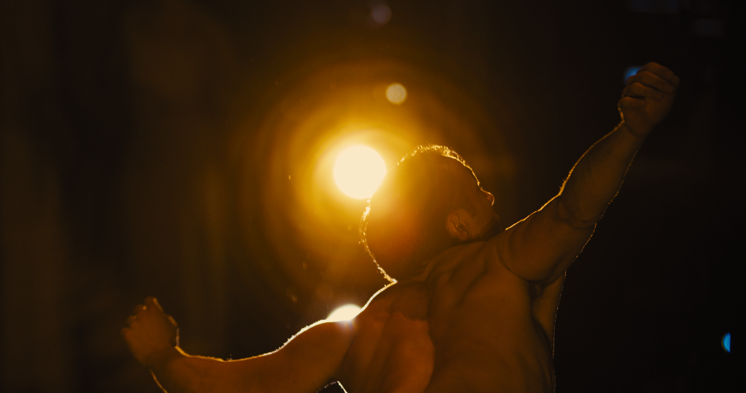 A statue of a muscular man with arms raised, backlit by a bright light, creating a silhouette effect in a dark background.