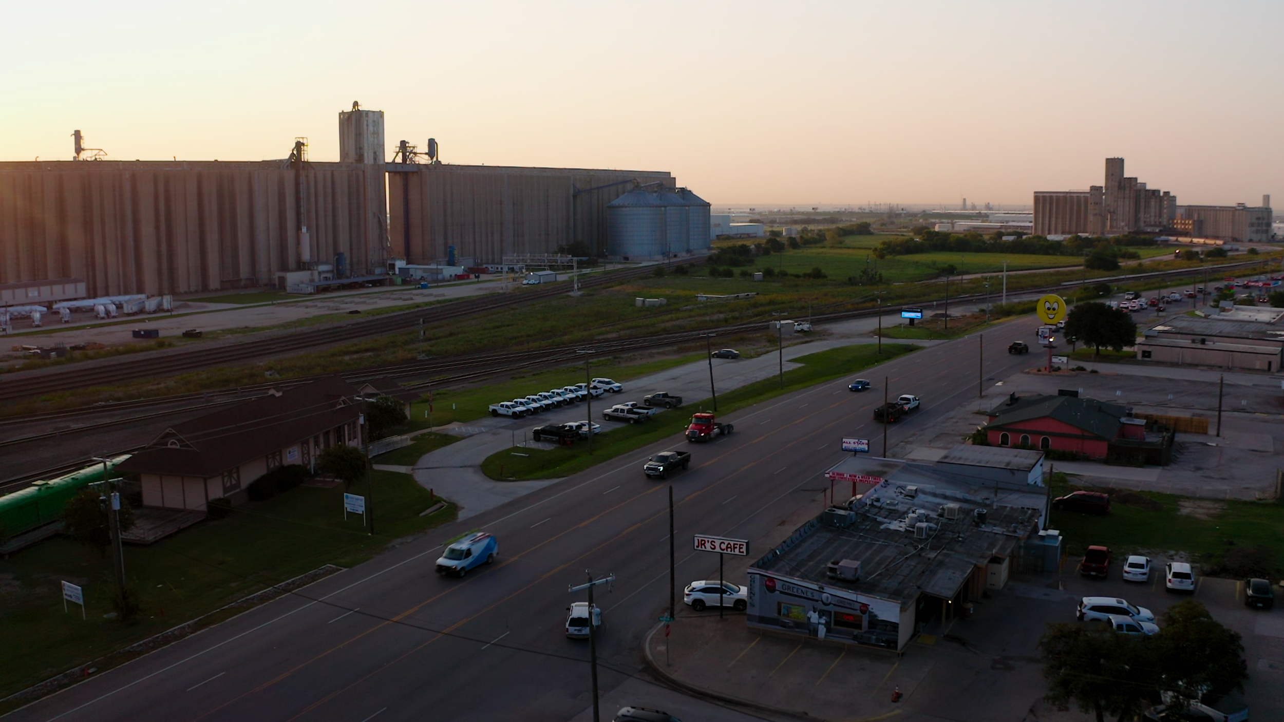 An urban scene at sunset with a street, parked cars, commercial buildings, railway tracks, grain silos, and distant industrial structures on the horizon.
