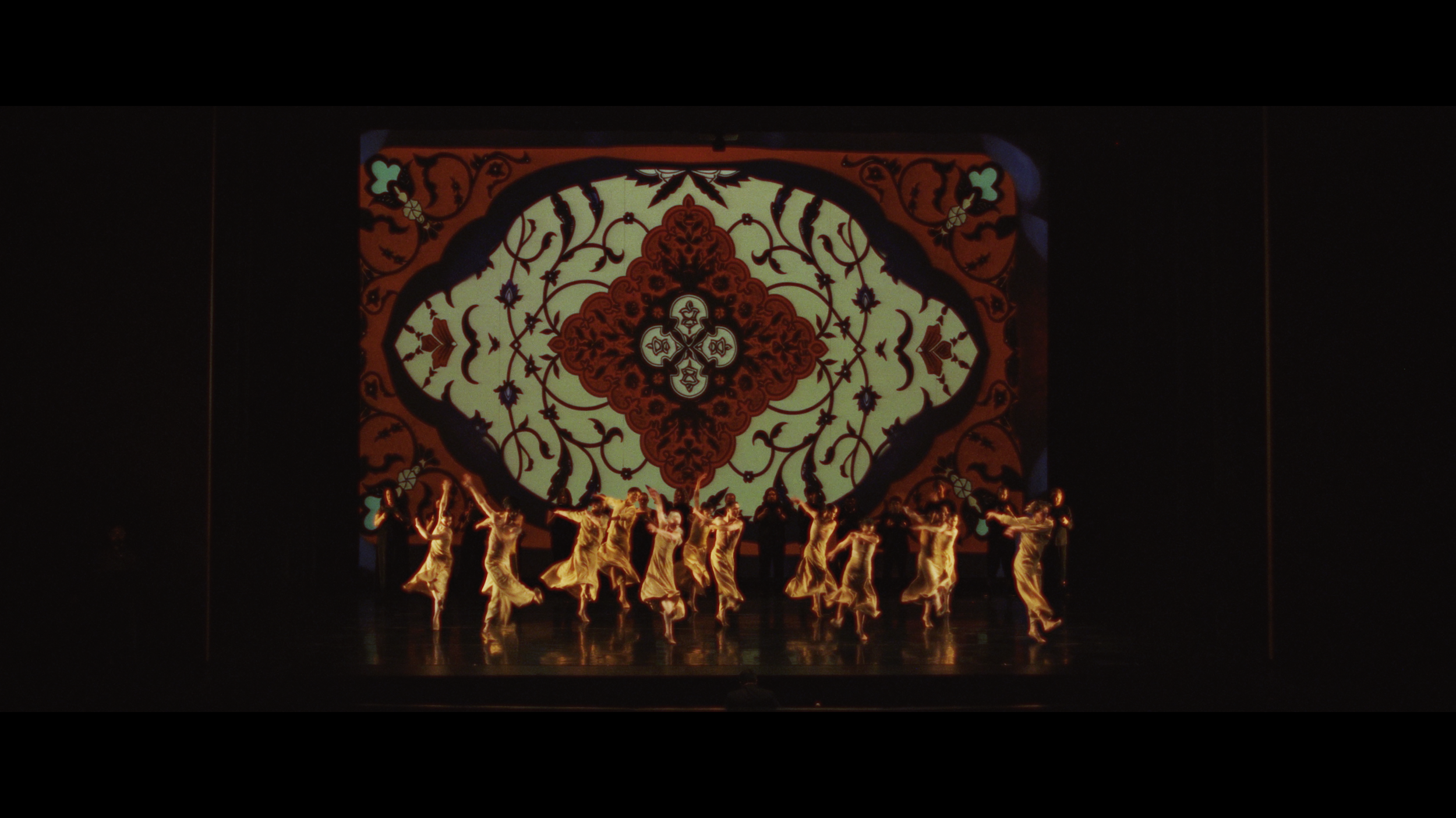 Dancers in gold costumes perform on stage in front of a large, ornate stained-glass window backdrop.