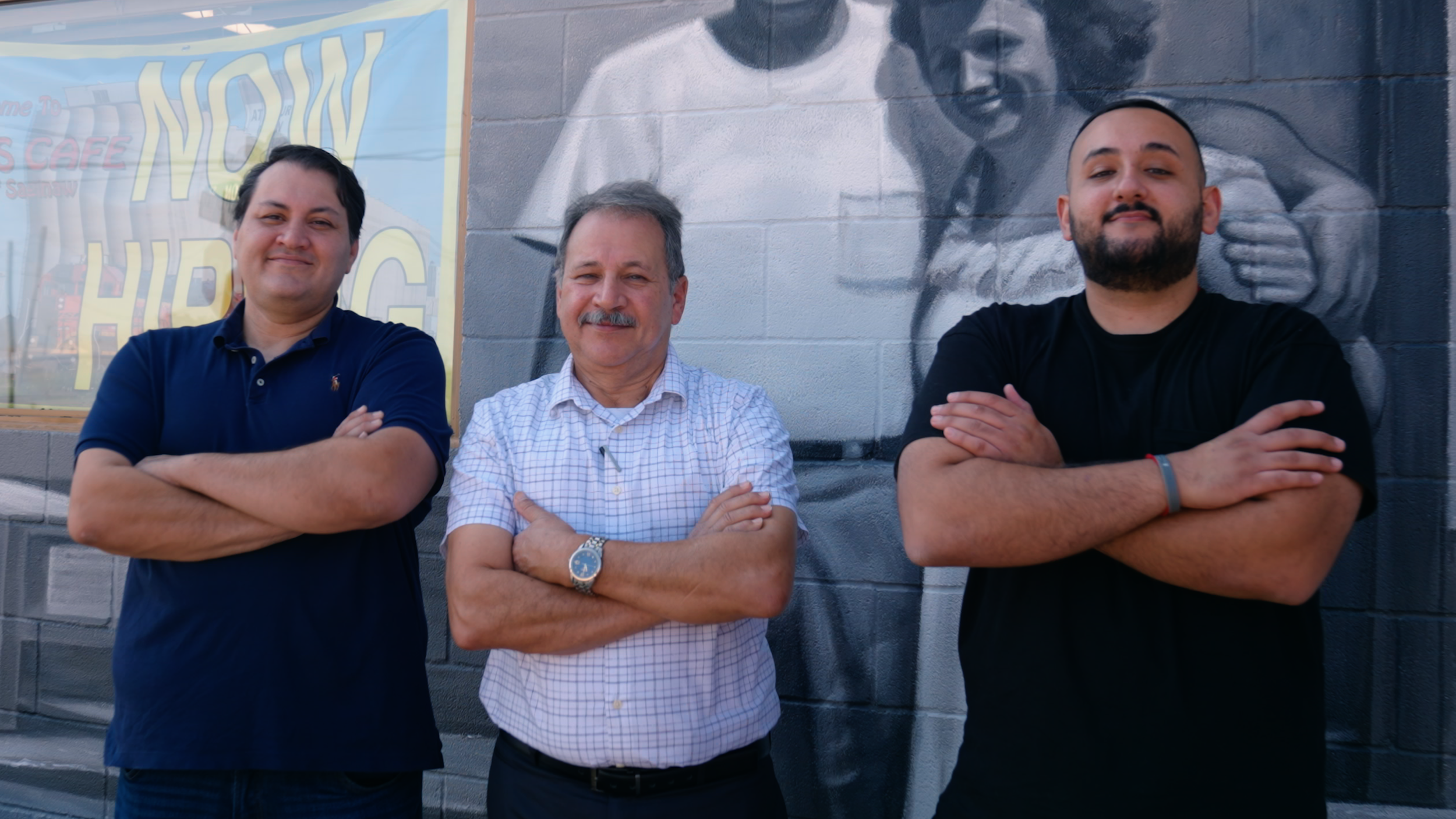 Three men standing in front of a mural on a brick wall with their arms crossed, smiling at the camera. Video production in Fort Worth, texas