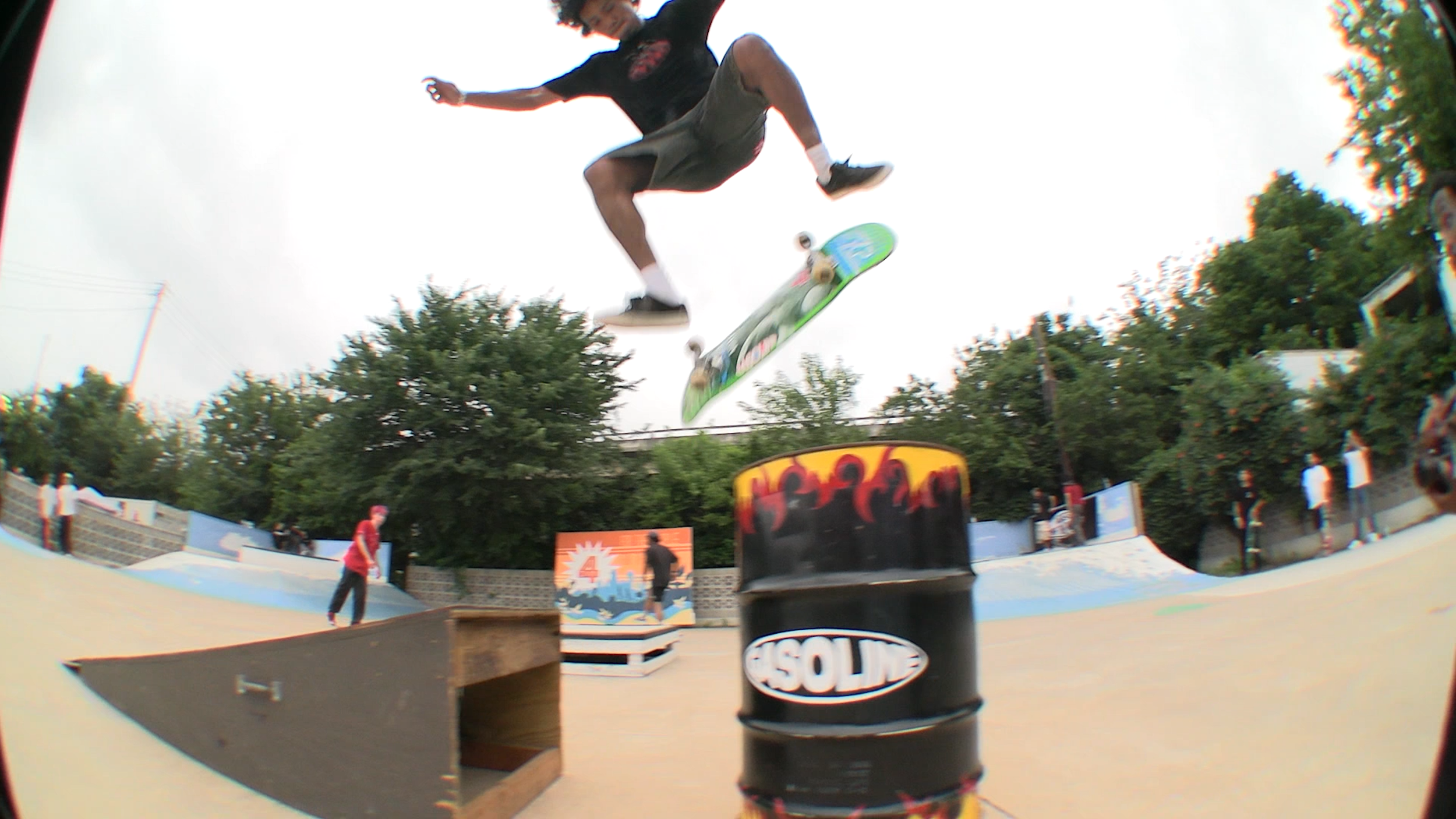 Skateboarder in mid-air performing a trick at skate park, with skateboarding obstacles and other skaters in the background under cloudy sky.