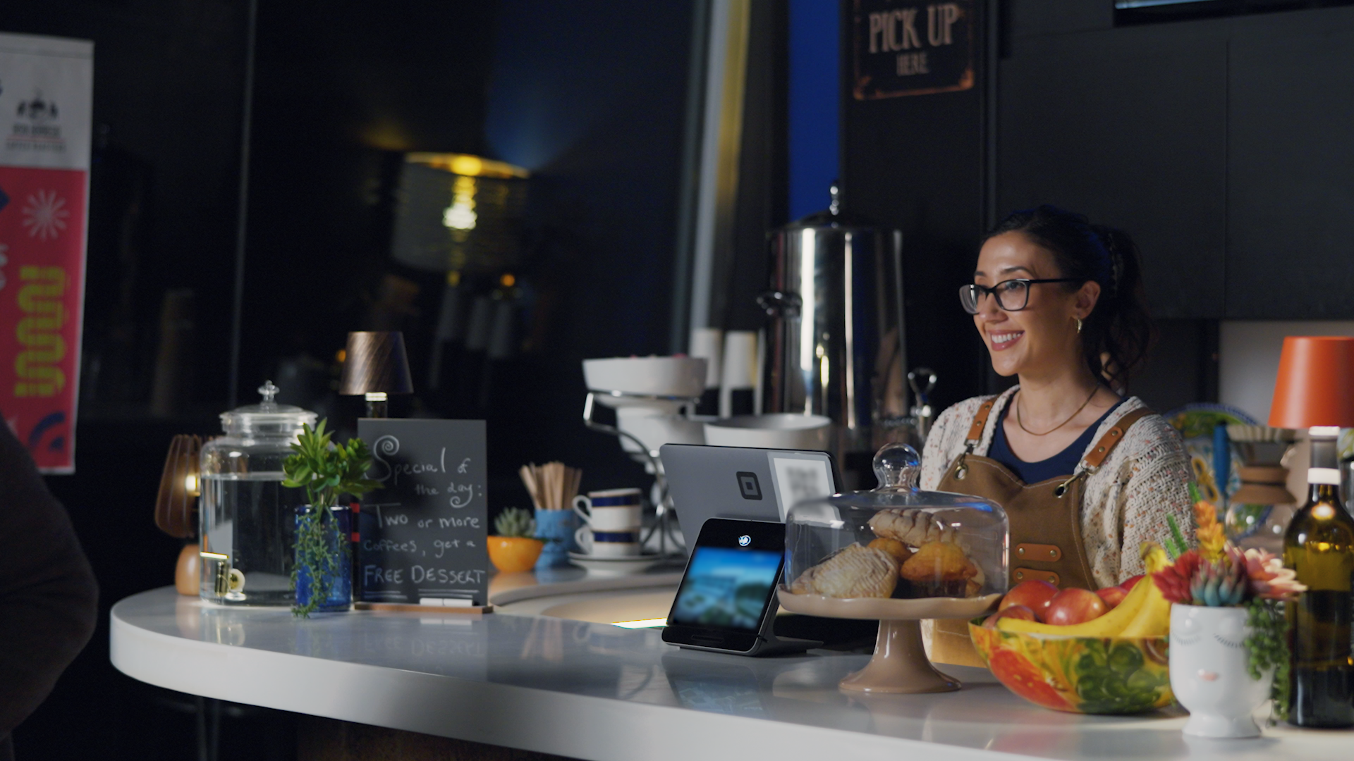 A smiling female barista with glasses and dark hair tied back, standing behind a coffee counter with baked goods, fruit, and a tip jar, in a modern cafe setting.