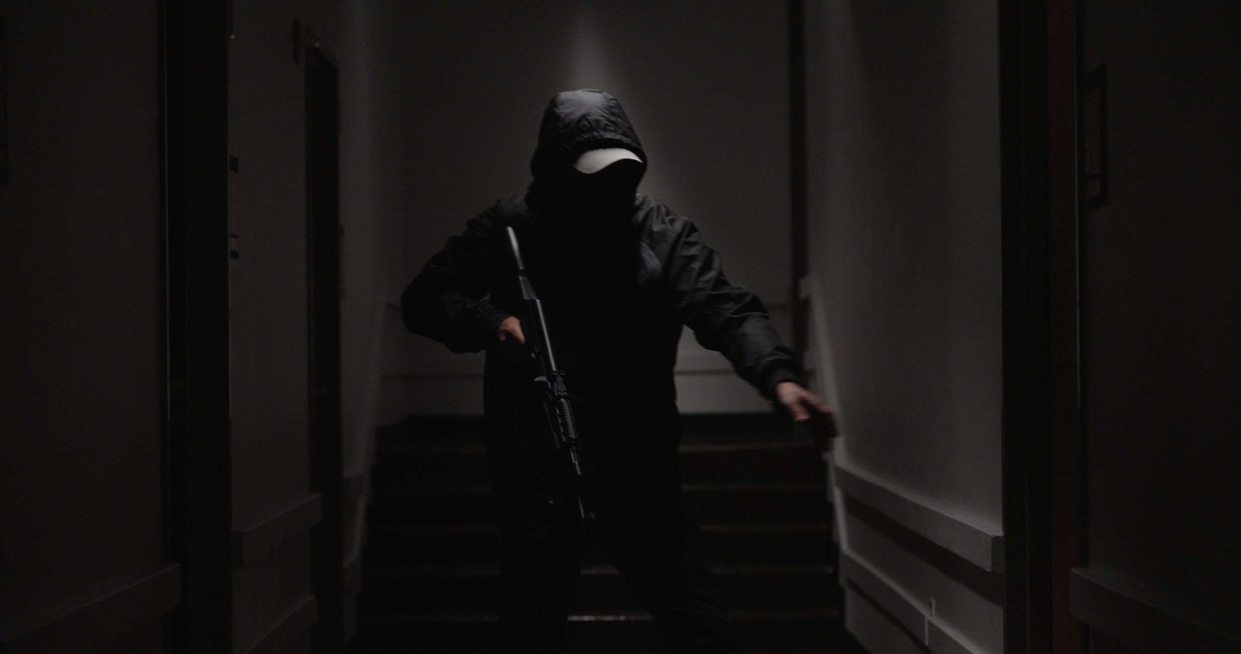 A person dressed in black with a hoodie and facemask, holding a gun, walking down a dark hallway.