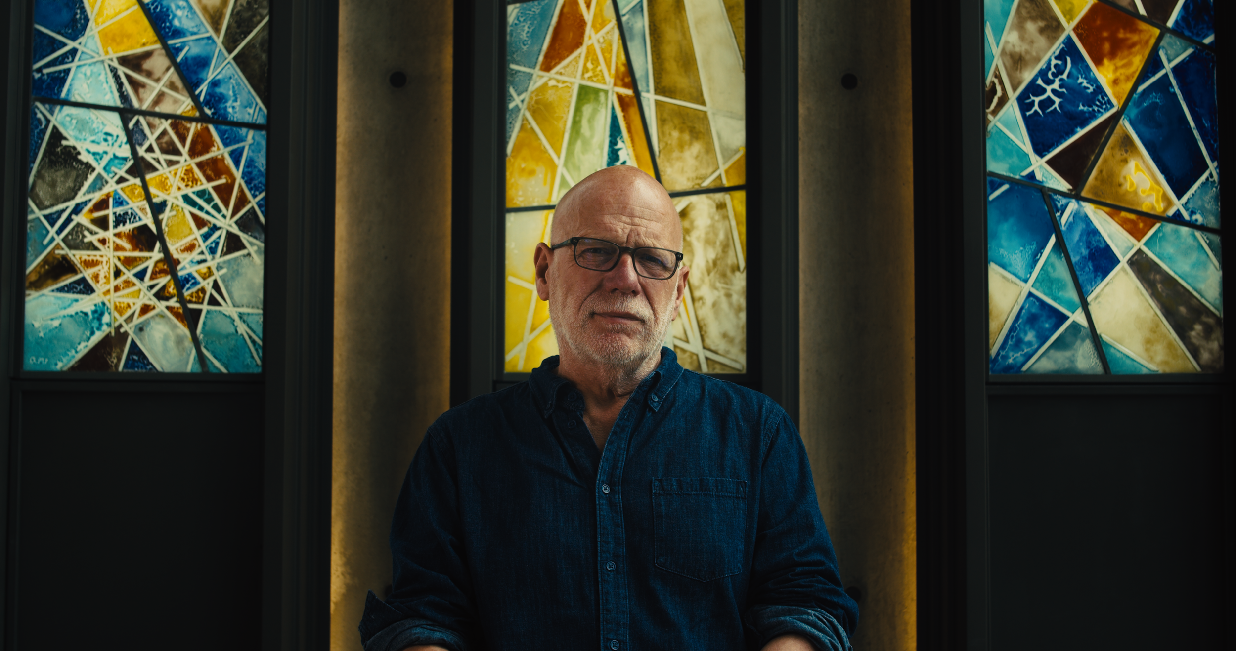 A man with glasses and a beard wearing a dark denim shirt in front of colorful stained glass windows. Documentary in Fort Worth texas.