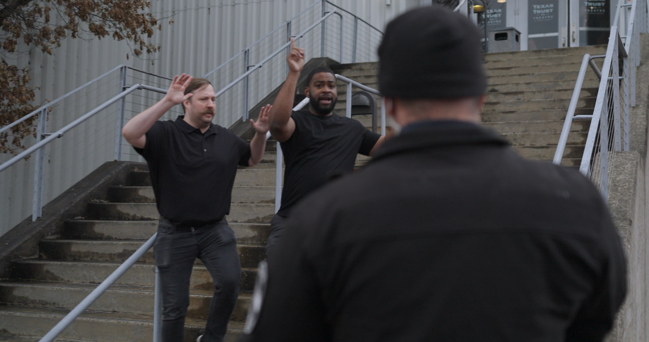 Two men with raised hands appearing to be in a confrontation or debate on outdoor stairs, while a third person in dark clothing faces them with his back to the camera.
