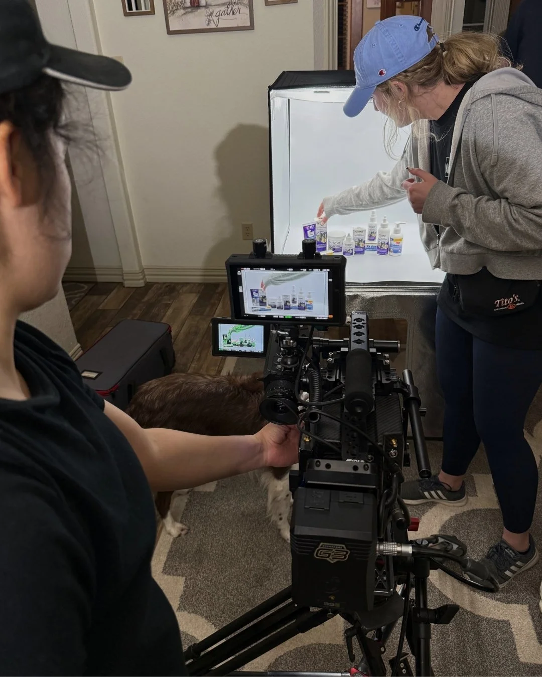 A woman in a light blue cap and gray hoodie is setting up bottles of medication or supplements behind a white backdrop, while a camera operator films her. The scene appears to be a product shoot or instructional video in a room with framed pictures o